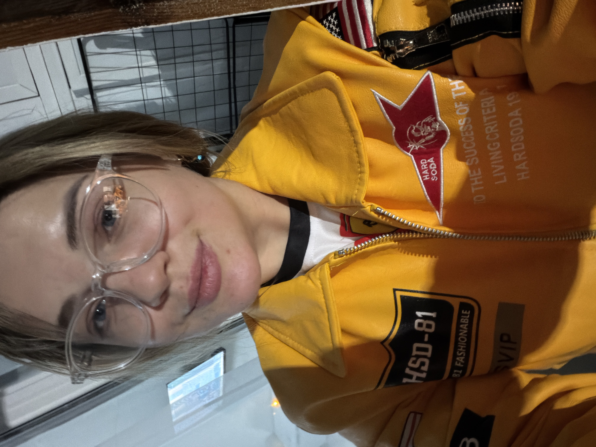 A woman wearing glasses and a yellow jacket with various logos poses indoors. The jacket has patches reading "HSD-81," "81 Fashionable," "VIP," and a "Hard Soda" logo. In the background, there's a grid pattern and other blurry objects, suggesting a shop or public space. The photograph does not show the city of Kazan, Russia.