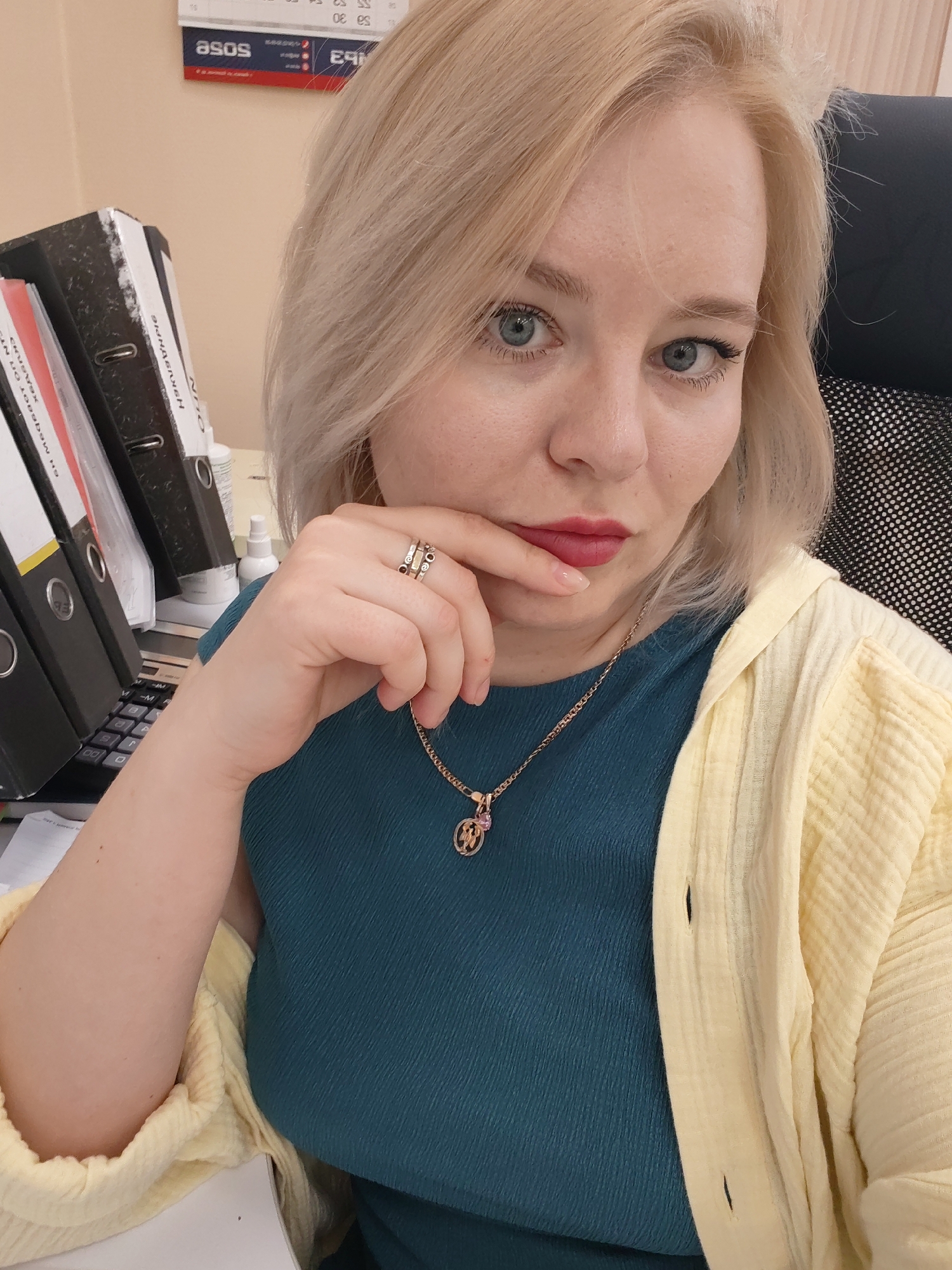 A woman with blonde hair and blue eyes sits at a desk in an office. She is wearing a teal top and a yellow cardigan. She has a gold necklace and rings on her fingers. Her right hand is touching her face. Behind her, there are files and a calculator on the desk. A calendar hangs on the wall, partially showing "ASOS" branding. The location is likely Izhevsk, Russia, given the presence of Cyrillic text on some of the files and the calendar.