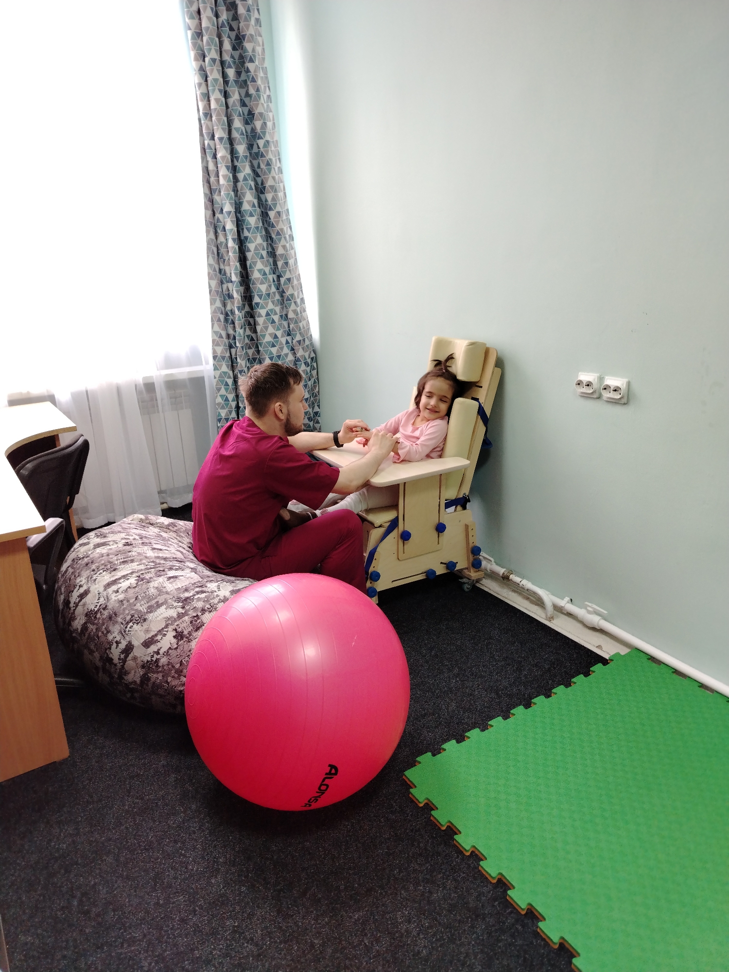 **Scene:** A brightly lit room in what appears to be a rehabilitation or therapy setting in Chaikovsky, Russia.
**People:** A young girl is seated in a specialized, adjustable wooden chair. She appears relaxed and possibly smiling. A man, presumably a therapist or caregiver, is seated on a large beanbag chair next to her, interacting with her hands.
**Objects:** In the foreground, a large pink exercise ball is prominently displayed. Behind the man, the beanbag chair has a mottled gray and brown pattern. The girl is seated in the wooden chair with blue straps. The room has a window with curtains patterned with blue and grey geometric shapes. There is a desk partially visible. On the wall, there are two electrical outlets. A section of green interlocking foam floor tiles is on the floor.
**Actions and Interactions:** The man is gently holding the girl's hands, possibly performing a therapeutic exercise or providing comfort. The girl appears receptive and calm.
**Environment:** The room is clean and well-lit, with a focus on therapeutic elements like the exercise ball and specialized chair. The black floor covering suggests it is a designated therapy area.