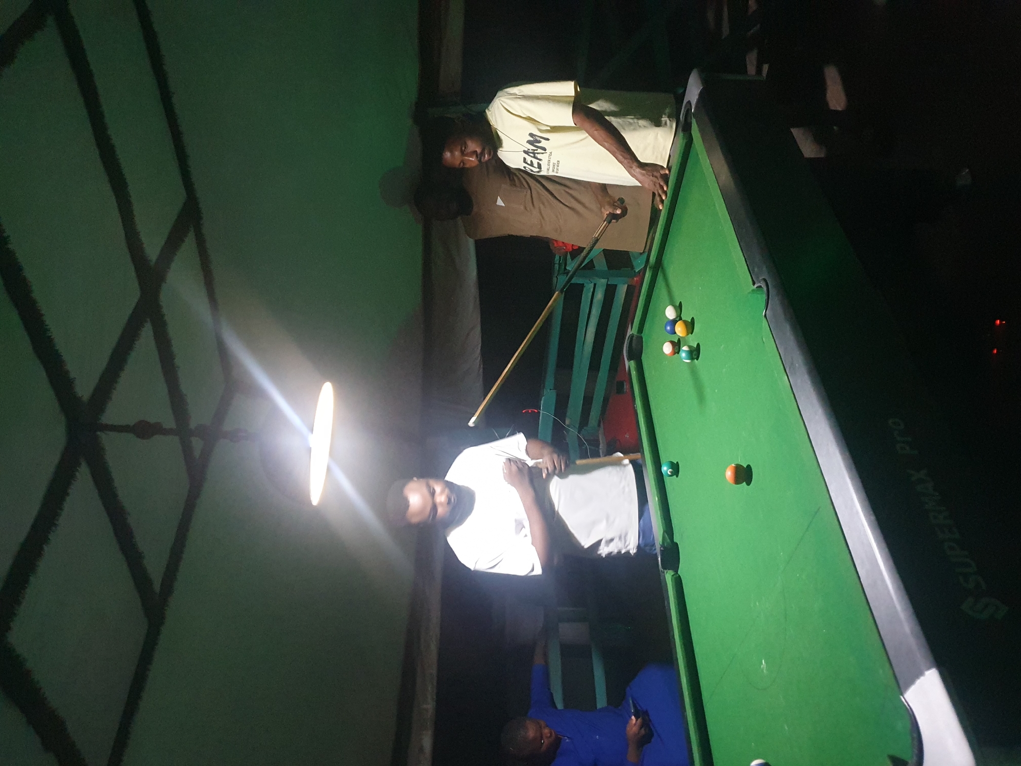 In Lokoja, Nigeria, a group of four individuals are gathered around a pool table inside a dimly lit, enclosed space, possibly a recreation area or bar, indicated by the presence of a pool table and a low ceiling with a grid pattern. The light source appears to be a single, bright overhead lamp, casting a warm glow on the scene.
On the left, a person in a blue garment is seated, looking down at a mobile device. In the center, a man in a white t-shirt is standing, holding a pool cue and seemingly contemplating his next move. To his right, another man, wearing a brown t-shirt, stands behind him. On the far right, a man in a yellow t-shirt with the word "REAM" printed on it, is also present, his hand resting near the pool table.
The pool table itself is green, with several colored balls scattered across its surface, including a green cue ball. The table has the brand name "SUPERMAX Pro" visible on its side. The background is dark, with some green structures that might be benches or railings. The overall atmosphere suggests a casual, recreational gathering.