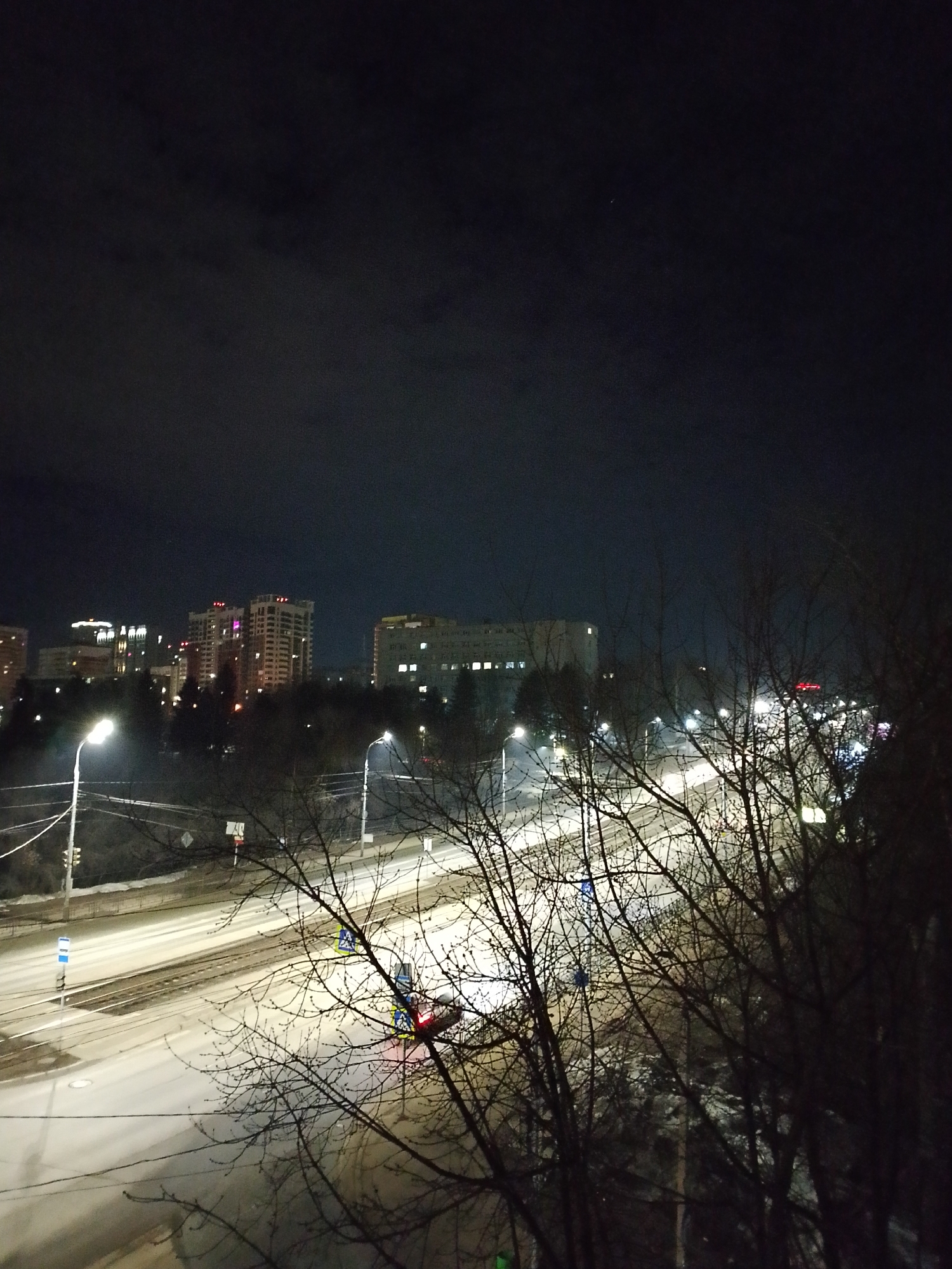 The image shows a nighttime urban scene in Izhevsk, Russia. A wide, snow-covered road is illuminated by streetlights, with a few vehicles visible, including a car with its tail lights illuminated. Bare trees dominate the foreground on the right side of the frame, partially obscuring the view. In the distance, there are buildings of varying heights, some with illuminated windows. The sky is dark and cloudy. The overall atmosphere is cold and dimly lit.