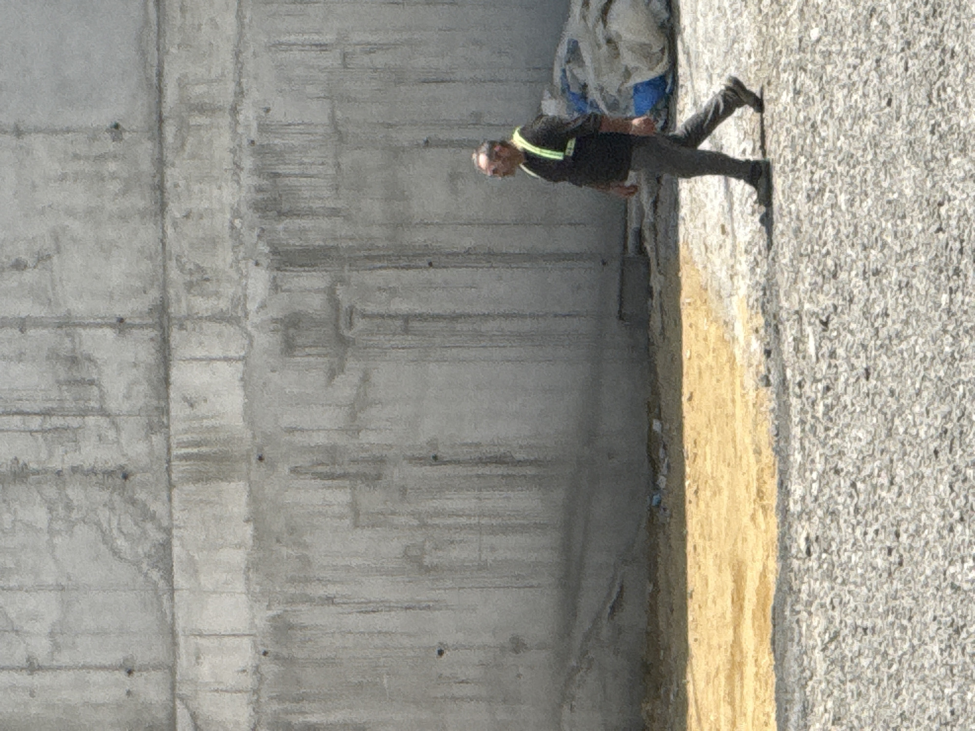 A man wearing a black long-sleeved shirt, gray pants, and a reflective yellow vest walks from right to left across a gravel and sand-covered area in front of a large, textured concrete wall. The concrete wall appears to be part of a construction site. There are some white and blue tarps or bags piled on the ground to the right of the man. The lighting suggests it is daytime. The scene appears to be an outdoor construction area in Tbilisi, Georgia.