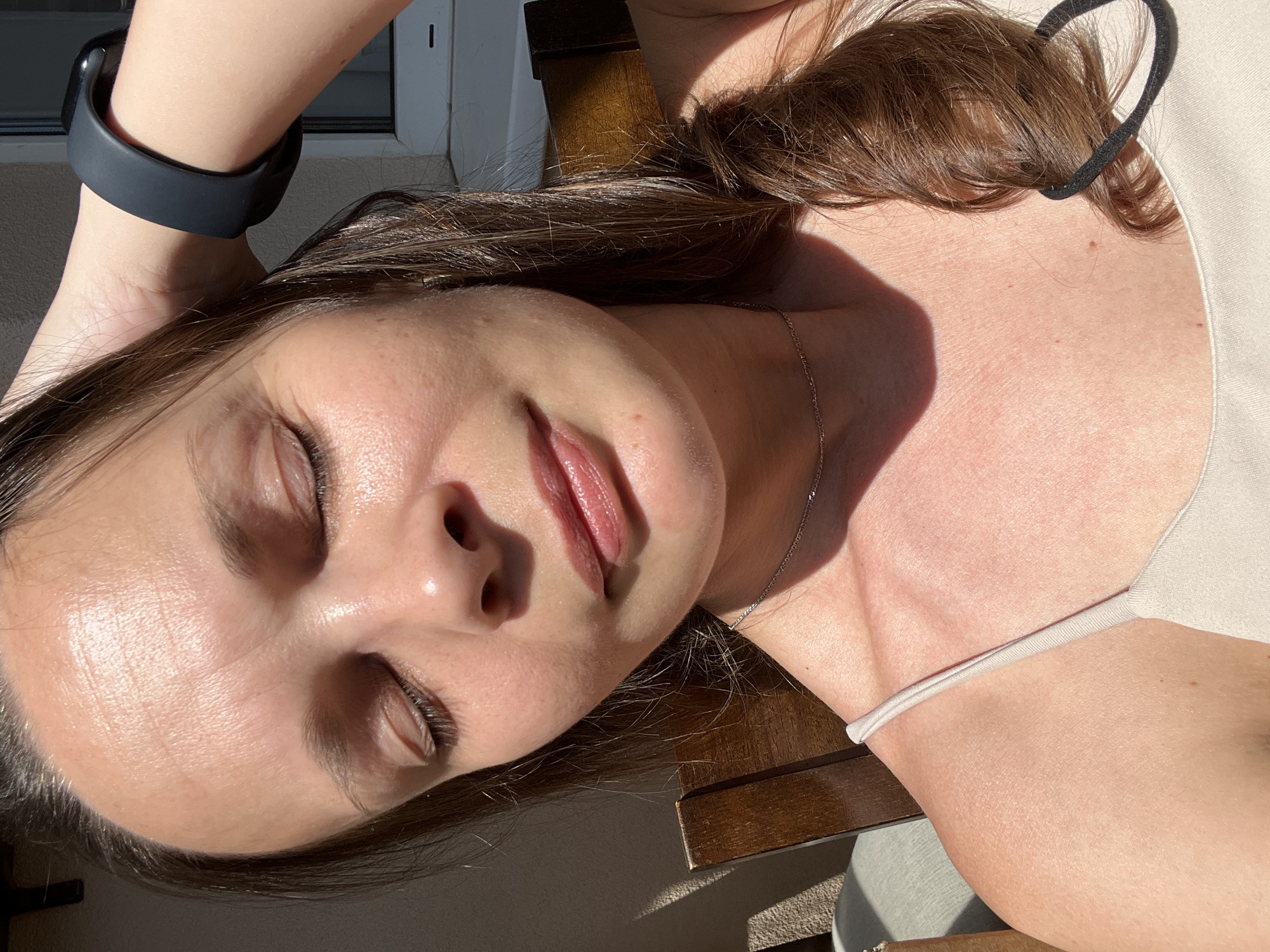 A medium shot, eye-level, close-up photograph captures a woman with her eyes closed and right hand raised to her head, touching her hair. She is looking slightly upwards and to her left, with her head tilted. Her lips are slightly parted, and her skin appears to be dewy and possibly illuminated by sunlight. She is wearing a light-colored tank top and a thin silver necklace. A black fitness tracker is visible on her left wrist. The background is blurred but suggests an outdoor setting with wooden furniture and natural light casting shadows. The image is a selfie, taken from a slightly low angle. There is no information to determine the location in Tolyatti, Russia.
