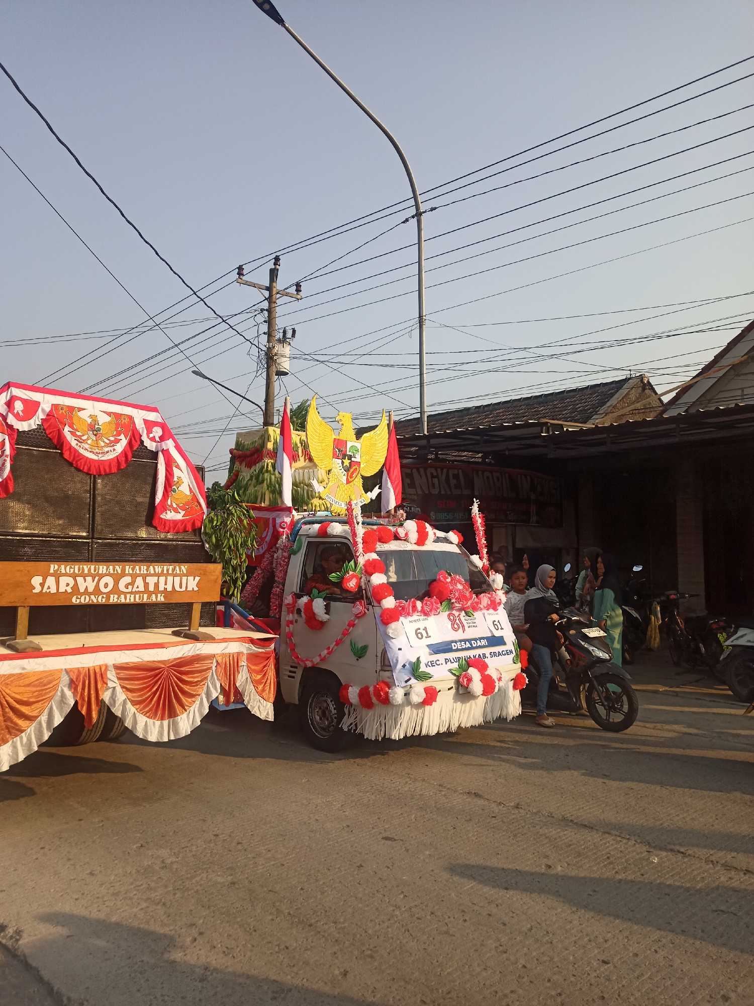 In the city of Ngrompongan, Indonesia, a decorated vehicle is participating in a parade. The vehicle, a white van, is adorned with red and white floral arrangements and banners. A large Garuda Pancasila emblem is prominently displayed on the side of the van, along with the text "DESA DARI KEC. PLUPURKAB. SRAGEN". To the left of the van, a trailer is set up with speakers and draped with red and white fabric, featuring the text "PAGUYUBAN KARAWITAN SARWO GATHUK GONG BAHULAK". Several people are gathered around the vehicle, some standing and others sitting. In the background, buildings and a utility pole with numerous wires are visible. The scene appears to be taking place outdoors during daylight hours, with shadows cast on the paved road.