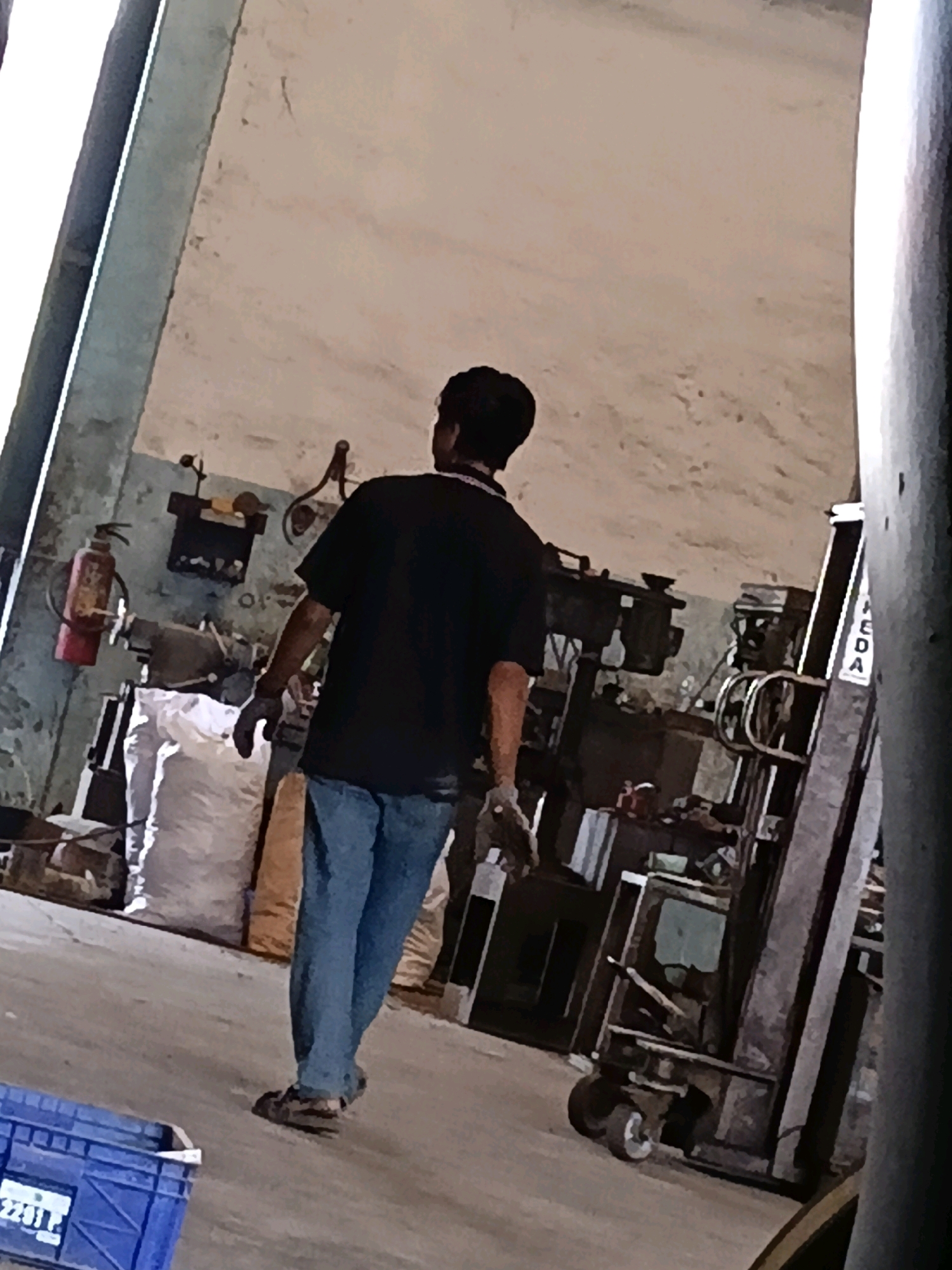 A person walks away from the camera in a dimly lit industrial interior. The person is wearing a black t-shirt, blue jeans, and dark shoes, with gloves on both hands. Behind the person are various pieces of industrial equipment and supplies, including a fire extinguisher mounted on a wall, large bags of material, a forklift, and what appears to be a workbench with tools. The floor is concrete, and the walls are a light color with visible texture. The scene suggests a workshop or storage area within a factory or warehouse. There is no specific indication of the city of Belimbing, Indonesia within the image itself.