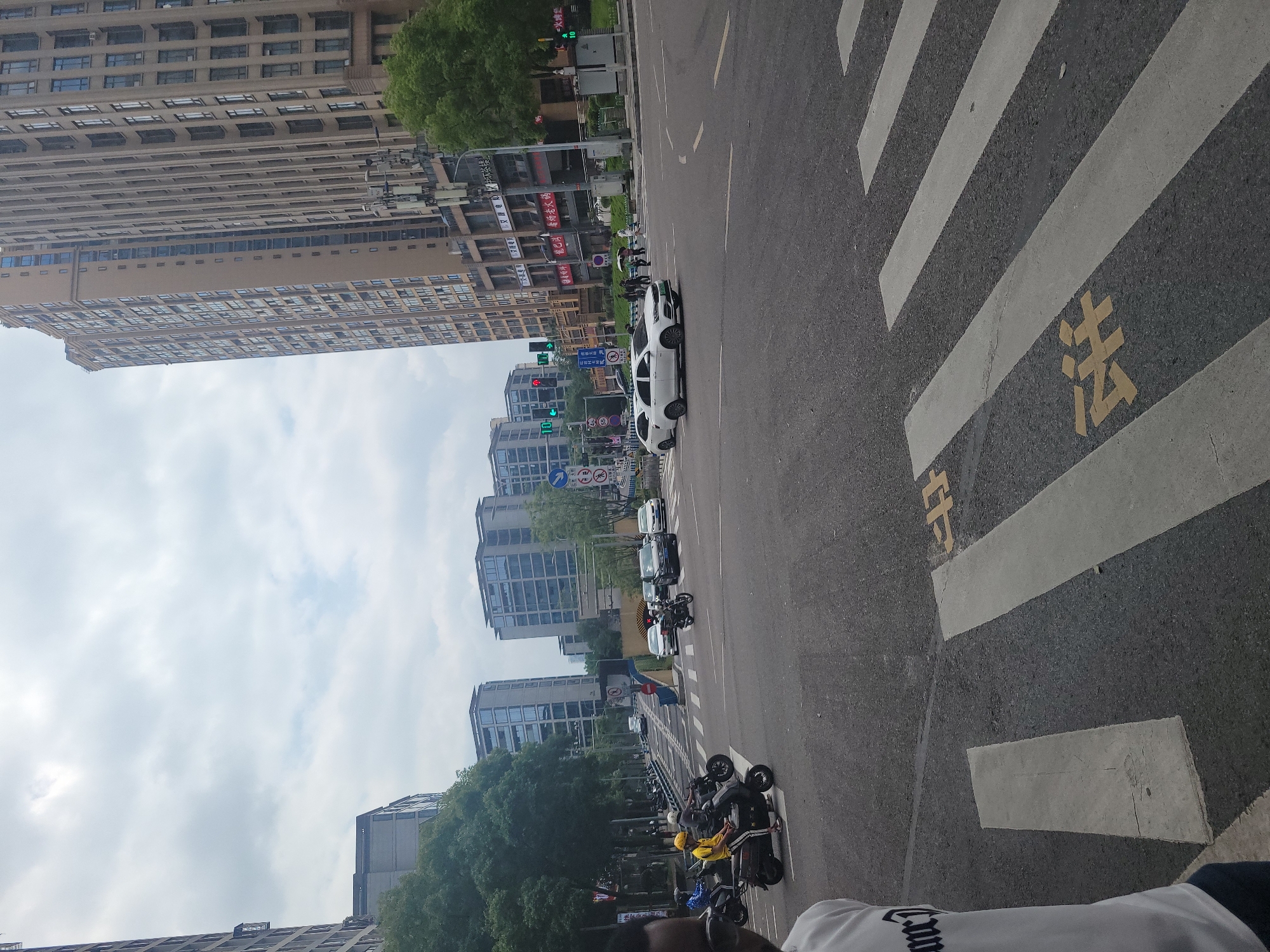 The image depicts a street scene in Yuzhong District, China, under an overcast sky. In the foreground, a pedestrian crossing with white stripes is visible on the asphalt road. Yellow markings indicating the crossing are present. To the left, the shoulder of a person wearing a white t-shirt with black lettering is captured.
The middle ground shows an intersection with several vehicles. Two individuals are riding on a scooter, one wearing a yellow helmet. Other vehicles, including motorcycles, cars, and a black SUV, are present. Traffic signals are visible, with one showing a red light. Pedestrians are crossing the street in the distance.
The background features several tall buildings, characteristic of an urban environment. One building on the right is a light brown, multi-story structure with numerous windows. To the left, other modern buildings with glass facades are visible. Trees line the street, and communication towers are also present. The overall scene suggests a typical day in a busy city.