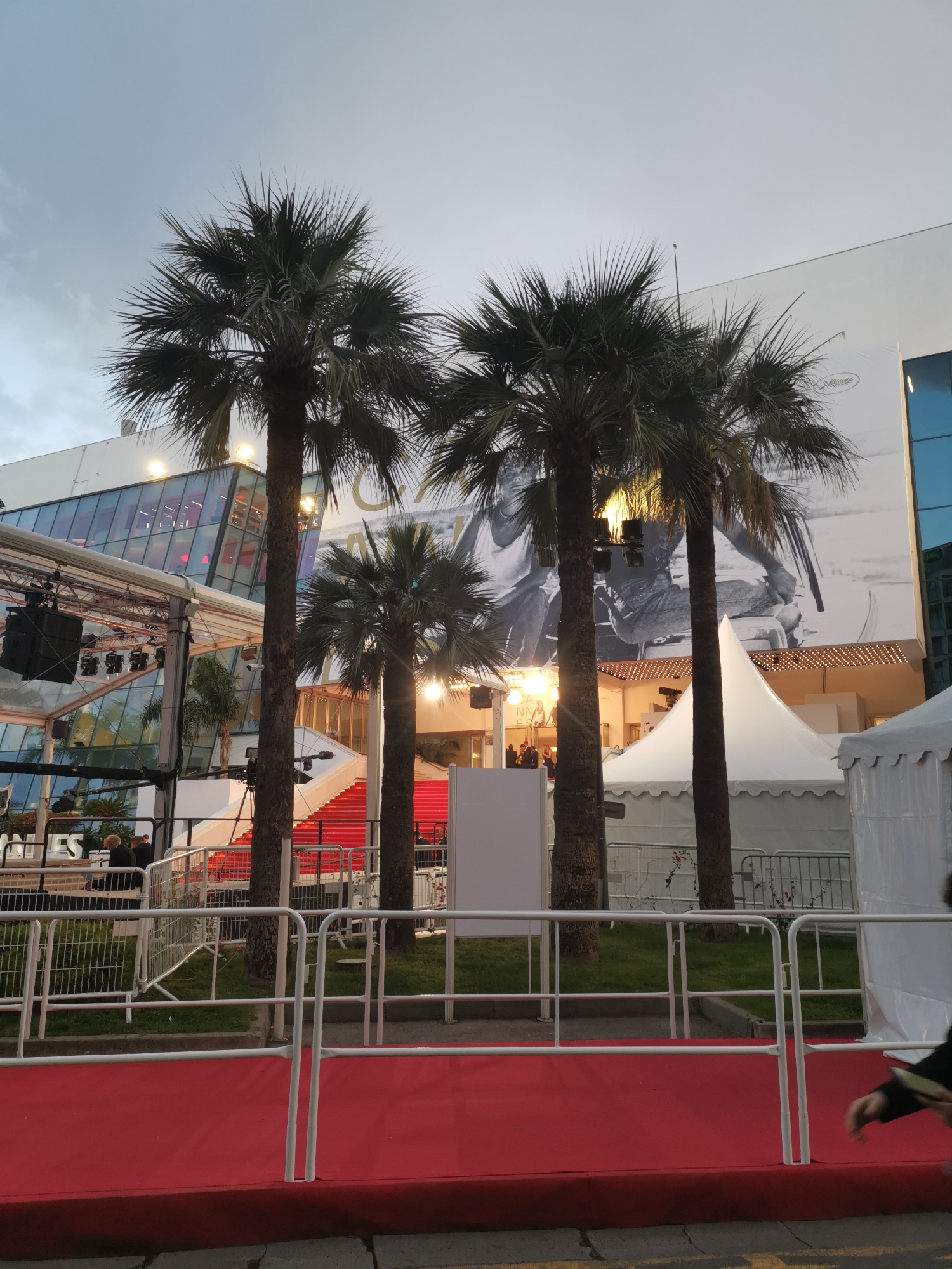 The image depicts a scene at the Cannes Film Festival in Cannes, France. The Palais des Festivals et des Congrès, the main venue, is visible in the background, adorned with a large black-and-white poster and prominent red steps. Several palm trees stand in front of the building. A red carpet is present in the foreground, bordered by white metal barriers. Tents and stage equipment are set up to the right and left, respectively. The sky is overcast, suggesting either early evening or late afternoon. There are several people in the background.