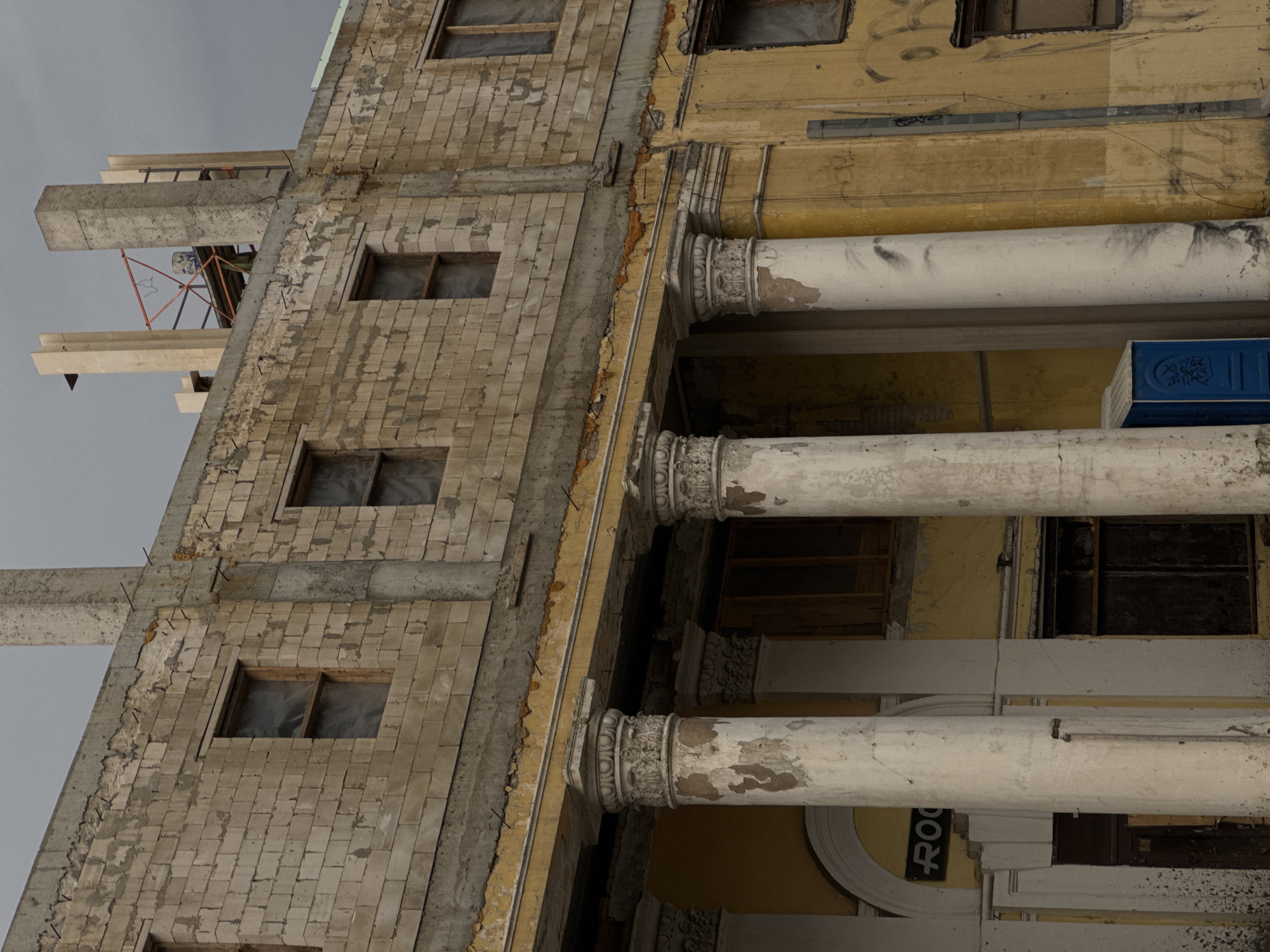 A weathered, pale yellow building with a pale brown brick facade stands under an overcast sky. The building's exterior shows signs of disrepair, with peeling paint and crumbling plaster. Large, classical-style columns with ornate capitals stand in front of the building, supporting a ledge. The columns are weathered and stained. Several windows on the upper floors are boarded up or covered with plastic. A blue portable toilet is visible to the right of the central columns, near the ground floor entrance. The word "ROO" is visible on a black sign above the entrance. The ground and lower walls are covered in graffiti. The overall scene suggests abandonment or ongoing construction/demolition.