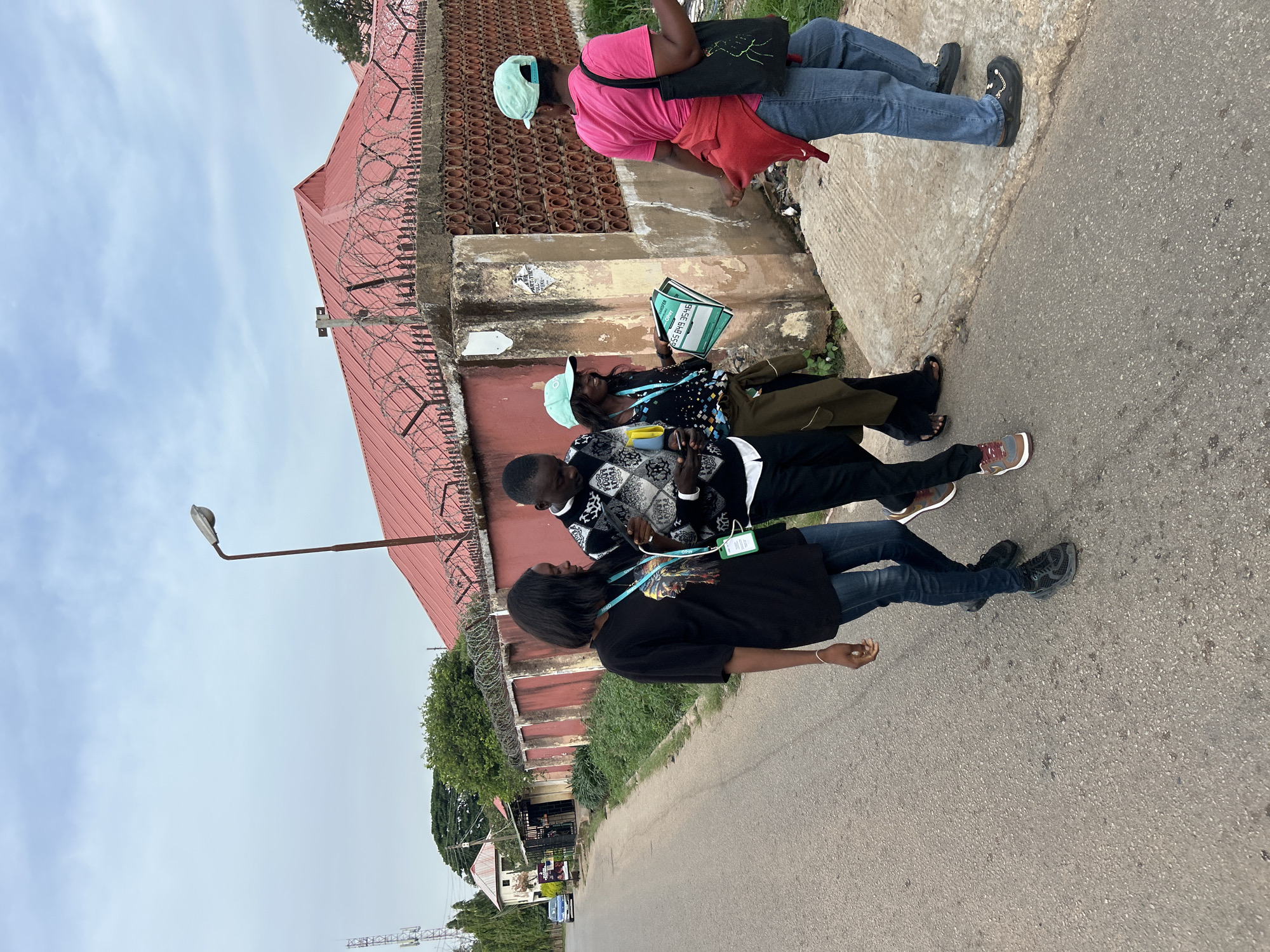 Three individuals stand on a sidewalk next to a road. A red-roofed building with barbed wire is visible behind them. The person closest to the camera wears a black t-shirt, jeans, and black shoes, and has a lanyard around their neck. The second person wears a black and white patterned sweater, khaki pants, and multi-colored shoes, and also has a lanyard. The third person wears a pink shirt, jeans, black shoes, a light blue hat, and a black bag. This image was taken in Jos, Nigeria.