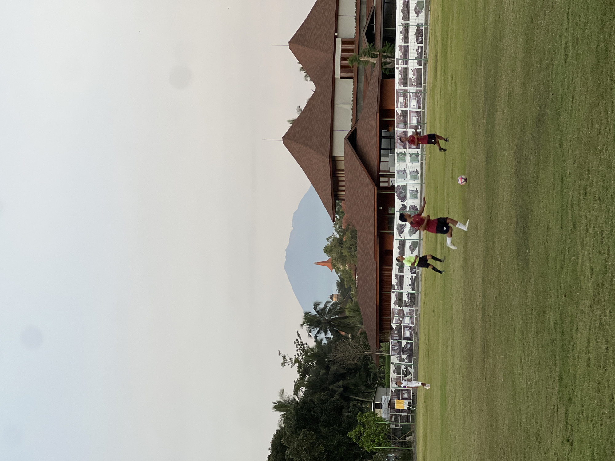 The image captures a scene in the Ubud District of Indonesia, featuring a grassy field adjacent to a building with multiple gabled roofs and a wall decorated with numerous framed images. Three individuals are actively engaged in playing soccer on the field. One player, wearing red and black attire with white socks, is in the midst of kicking a soccer ball. Another player, also dressed in red and black, is positioned nearby. A third player, wearing a lime green shirt and black shorts, is also visible. In the background, a mountain and a temple structure can be seen. The environment is green and the sky is overcast.
