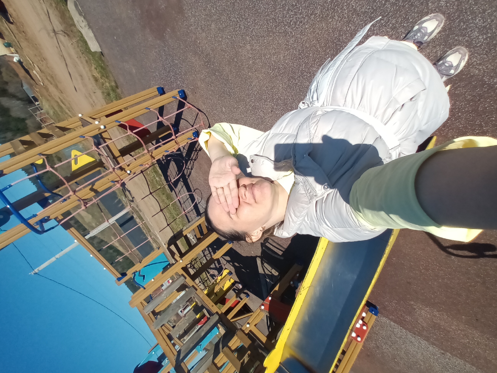 A medium shot taken at ground level depicts a woman standing adjacent to a children's playground structure, located in Ольховка, Russia. The playground equipment consists of a wooden frame with net climbing elements and a yellow slide. The woman is dressed in a light-colored jacket and shoes. Her hand is raised to shield her eyes from the sun. The background shows a dirt area and glimpses of other urban elements.