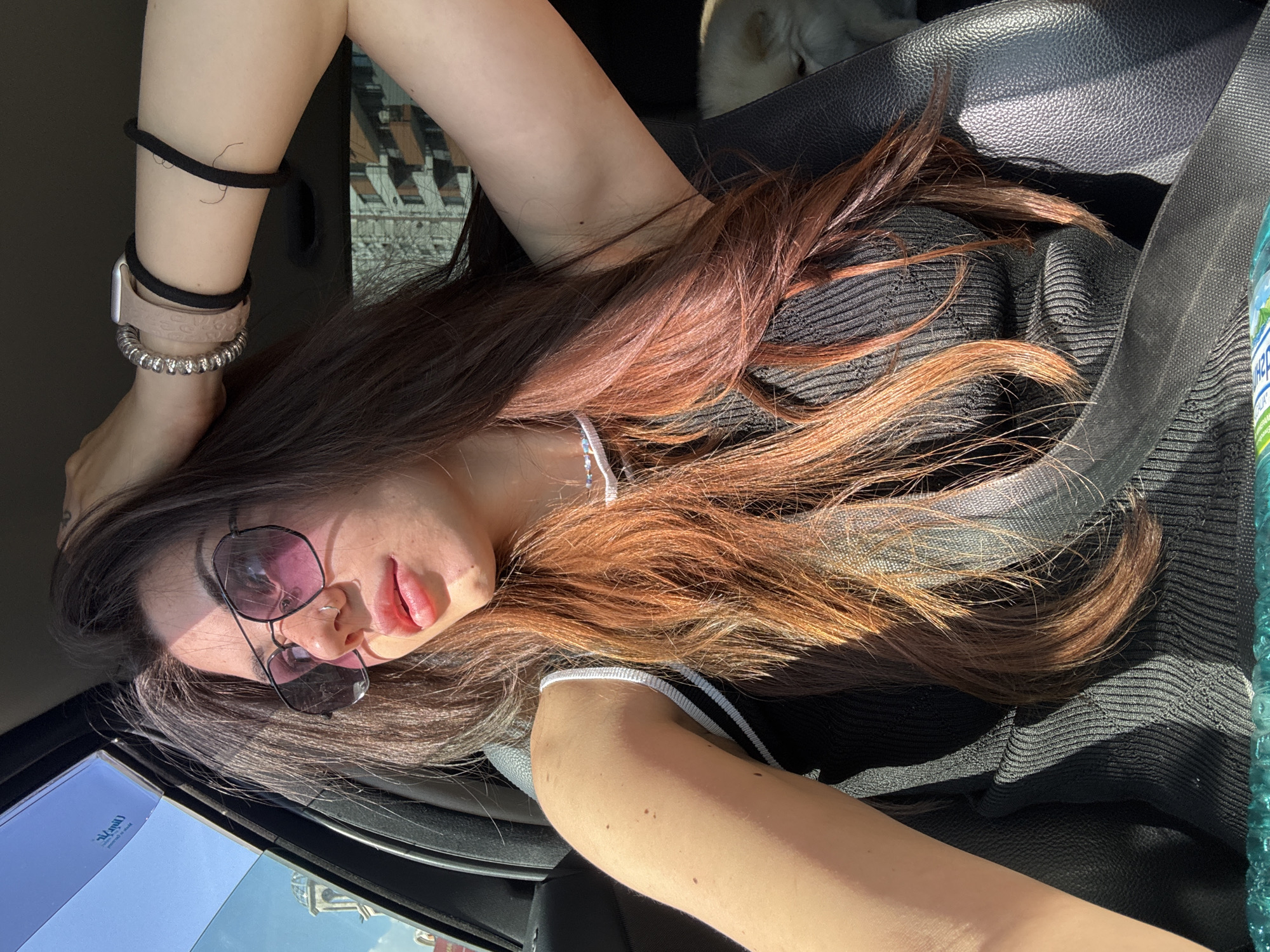 A young woman with long, dark auburn hair is pictured from the chest up, sitting in the passenger seat of a car. She is wearing sunglasses with pink lenses, a black ribbed tank top, and a silver beaded bracelet and a watch on her left wrist, along with a black hair tie. Her right arm is raised and resting on her head, obscuring part of her face. Sunlight streams in from the side window, casting shadows across her face and hair. In the background, a small white dog is visible in the passenger seat behind her. The exterior of buildings and a building with several balconies can be seen through the car window, suggesting an urban setting. The image appears to have been taken during the daytime.