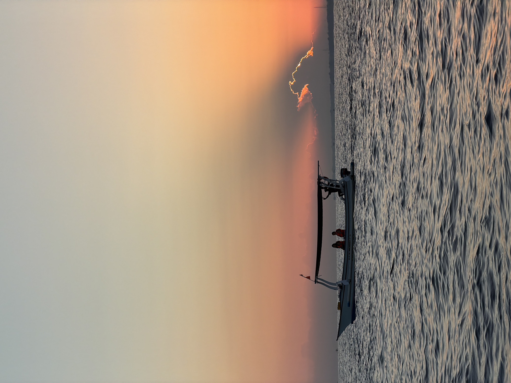 An eye-level medium shot captures a small motorboat with a canopy sailing on the ocean at sunset. The boat carries at least three people: two passengers wearing orange life vests are seated, and a third person, presumably the captain, stands near the stern. The sky exhibits a gradient of colors, transitioning from a pale blue at the top to soft orange and pink hues near the horizon, where the sun is setting behind a cloud. Sunrays emanate from behind the cloud, creating a dramatic visual effect. The ocean water is a deep blue with visible ripples, reflecting the colors of the sky. In the distance, a faint silhouette of land and possibly buildings can be discerned. The overall scene is tranquil and evokes a sense of peace at the end of the day. The location is noted as Unknown, Indonesia.