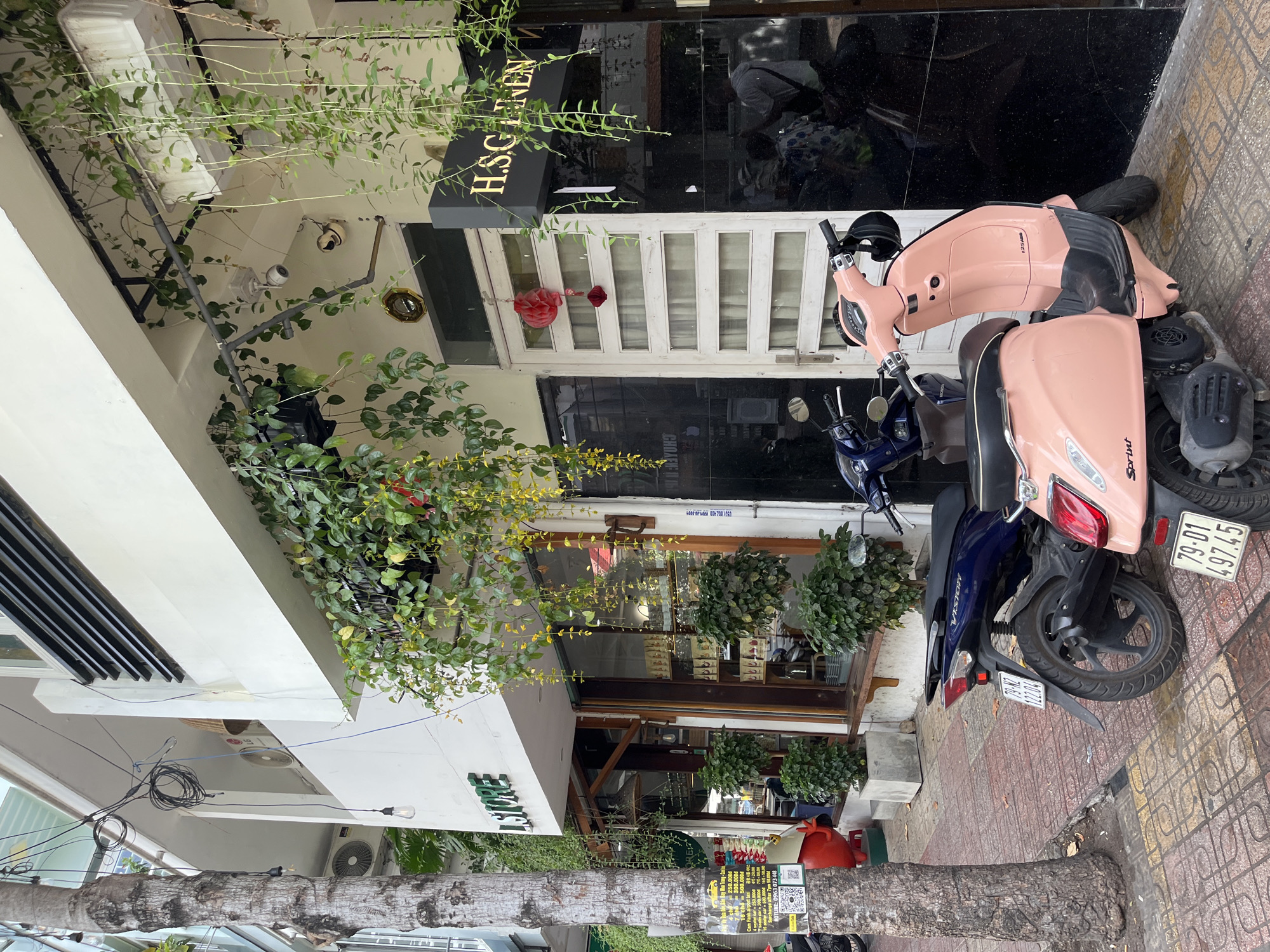 The image depicts a street scene in Nha Trang, Vietnam, likely in a commercial area given the storefronts and parked vehicles. In the foreground, two scooters are parked on a patterned tile sidewalk. The scooter on the right is a light pink Vespa-style scooter with "Sprint" visible on its side, and the license plate reads "79-D1 497.45". The scooter on the left is dark blue, with the model name "VISION" visible on its rear. Its license plate reads "79-NZ 122.04".
Behind the scooters, there is a building with multiple storefronts. The left storefront has signage that reads "L STORE" in green lettering. Its windows display various products on shelves. To the right of "L STORE" is a white door with multiple horizontal panes. Further to the right, a black facade building displays a dark sign with gold lettering that reads "H.S.G LINEN". Hanging from the building's overhangs are lush green plants and vines, some with yellow flowers. A red decorative lantern hangs from the white door.
The sidewalk is paved with square tiles in varying shades of red and yellow. A large tree trunk stands on the left side of the image, with electrical wires and some hanging light fixtures visible above. Part of a red, cartoonish figure is visible near the tree. In the background, through the windows of "L STORE", a few potted green plants can be seen. Reflections in the black facade of the building suggest the presence of people and possibly other vehicles. An air conditioning unit is visible on the exterior wall of the building.