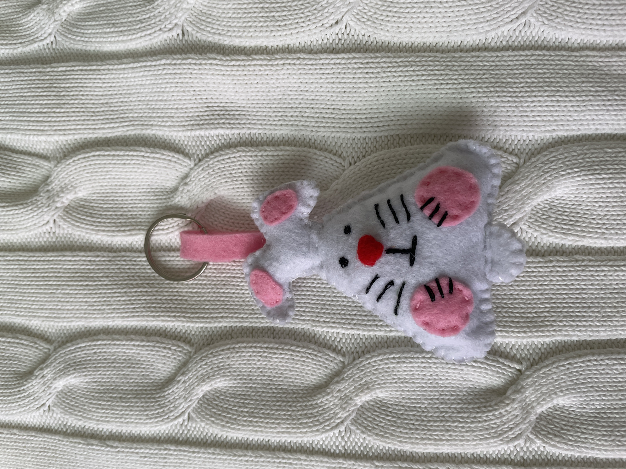 The image shows a handmade felt keychain of a white bunny with pink details on a white cable-knit sweater. The bunny has a red nose and black whiskers. The keychain has a silver ring and a pink felt loop. The background is the texture of the sweater. The location is Лесной Пейзаж Кп (шарапово, Russia is not evident from the image.