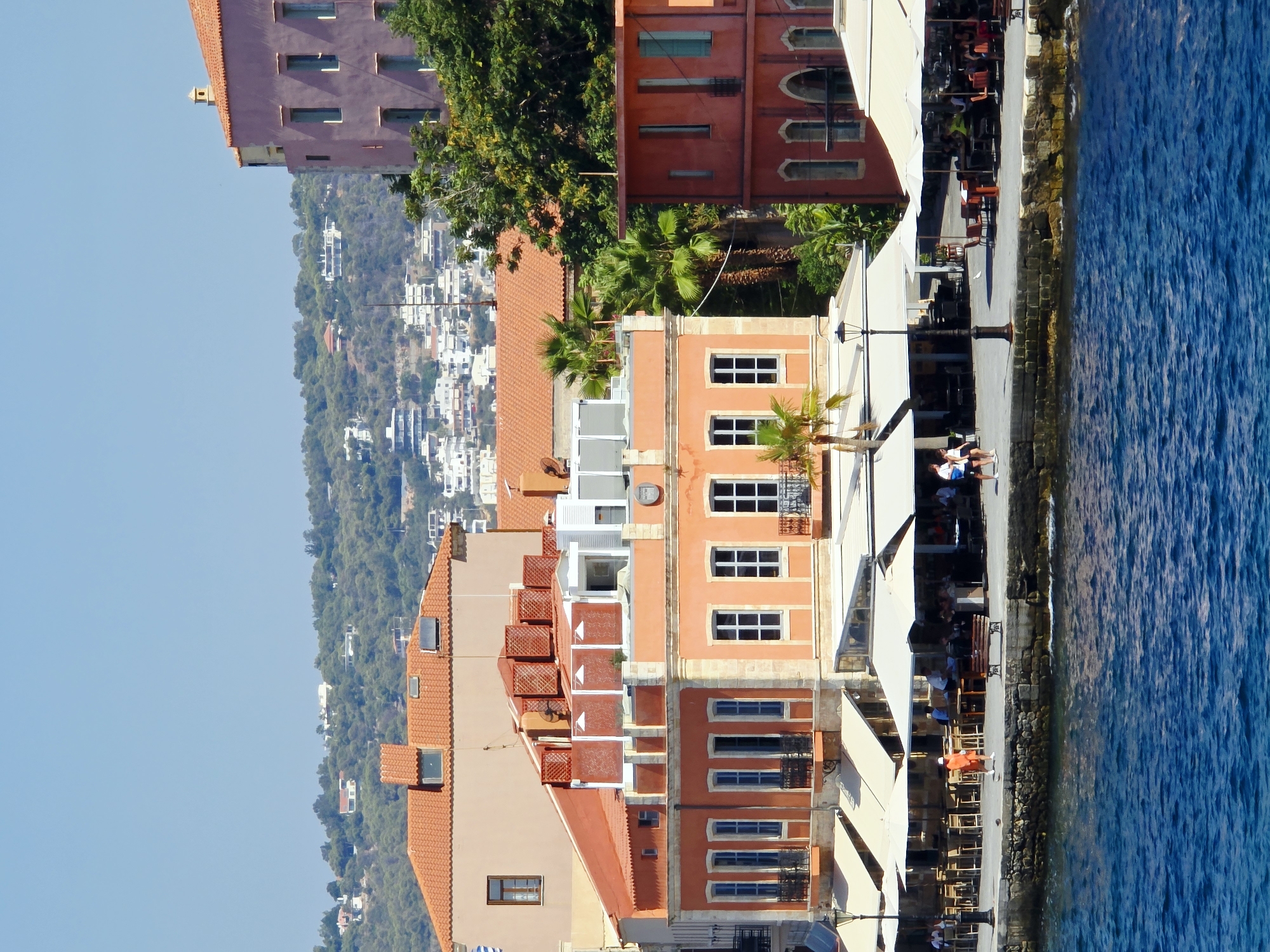The image depicts a waterfront scene in Chania, Greece, specifically likely the Venetian Harbor. In the foreground, the bright blue water of the sea gently laps against a stone embankment. Lining the embankment are several buildings with distinctive architecture. The main building in the center is painted in warm orange and peach tones, featuring multiple windows with balconies and awnings extended over outdoor seating areas. Adjacent to this, to the right, is a narrower, red-toned building with arched windows on its lower level and rectangular windows on the upper levels. Further to the left, a larger building with an orange tiled roof and a lighter-colored facade is partially visible. Behind these structures, a lush green hillside is covered with trees and dotted with houses, suggesting the city extends inland and upwards. The sky above is a clear, bright blue. Along the waterfront promenade, several people are visible. Some are seated at tables outside the cafes and restaurants, while others are walking or standing. The atmosphere appears to be a sunny, active day at a popular tourist destination.