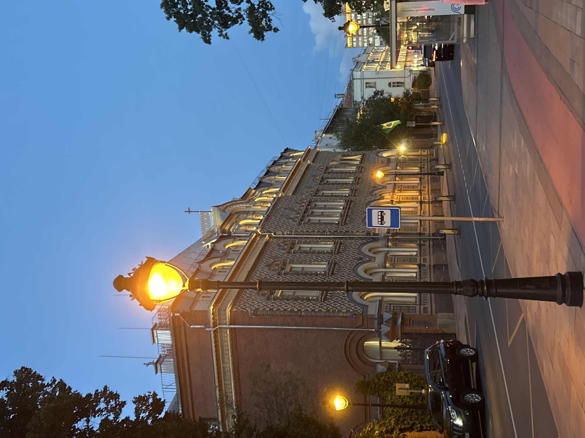 The image depicts a street scene in a city at dusk. A prominent, ornate black lamppost with an illuminated yellow lamp stands in the foreground, casting a warm glow on the patterned brick building to its left. This building features multiple arched windows and decorative stonework. Across the street, a bus stop shelter is visible with a sign indicating a bus stop. Several cars are parked or driving along the road, including a black SUV near the left side of the frame and another vehicle further down the street. The road is paved with red and grey bricks, with painted lane markings. A Brazilian flag is displayed on a pole between the brick building and the road. Trees line the street, and the sky is a deep blue, transitioning from day to night.