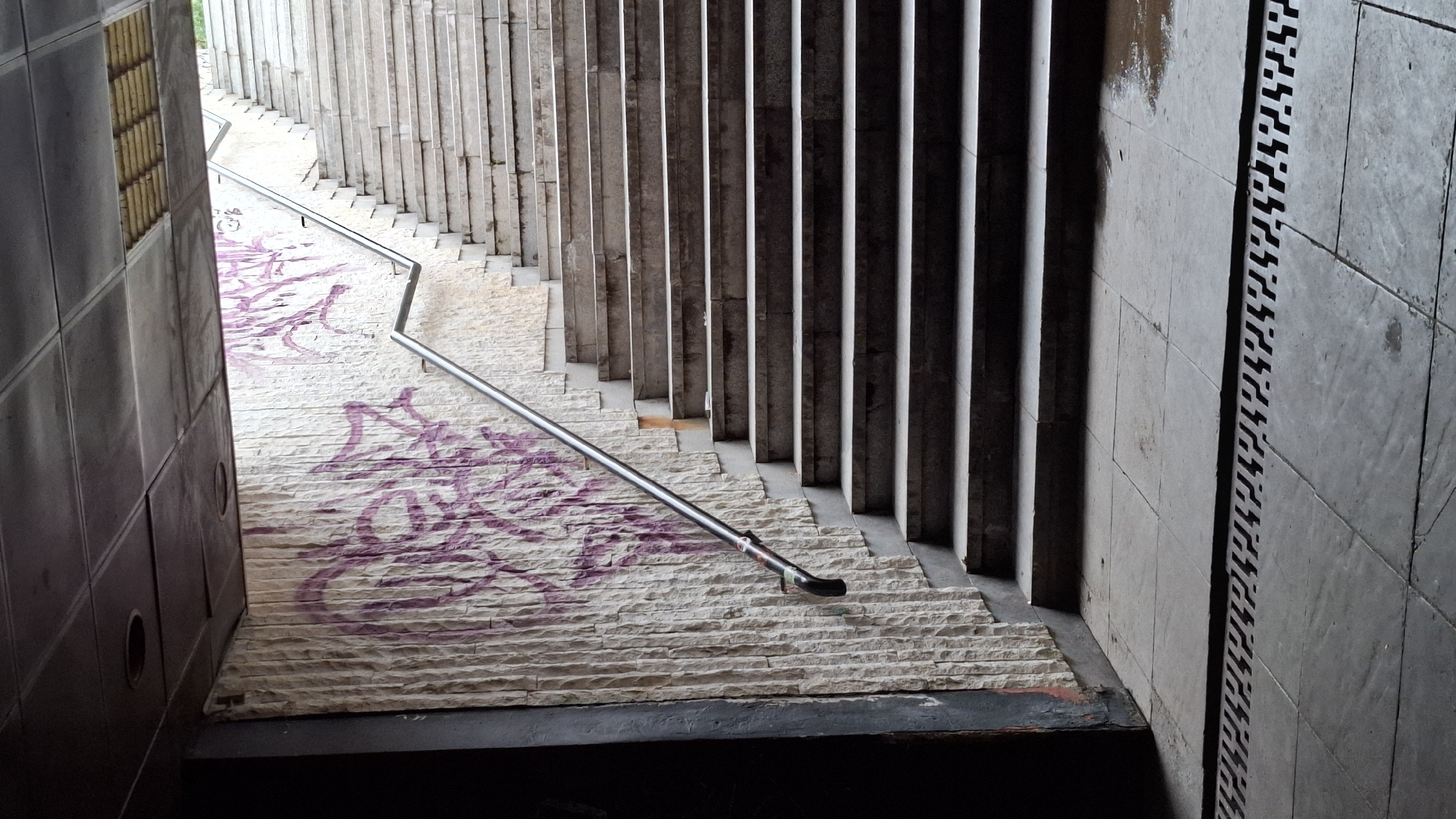 The image shows an outdoor staircase in an urban environment, possibly a pedestrian underpass. The staircase is made of concrete or stone steps and has a metal handrail on the right side. The wall to the left of the stairs is covered in textured stone and features purple graffiti. The ground at the base of the stairs is paved with stone tiles, with a drainage grate running across the foreground. The lighting suggests it is daytime, with sunlight entering from above and illuminating the top of the stairs. The scene is devoid of people. The architectural style and the presence of graffiti are consistent with many underpasses found in Sofia, Bulgaria, a city known for its extensive network of pedestrian underpasses and public art.