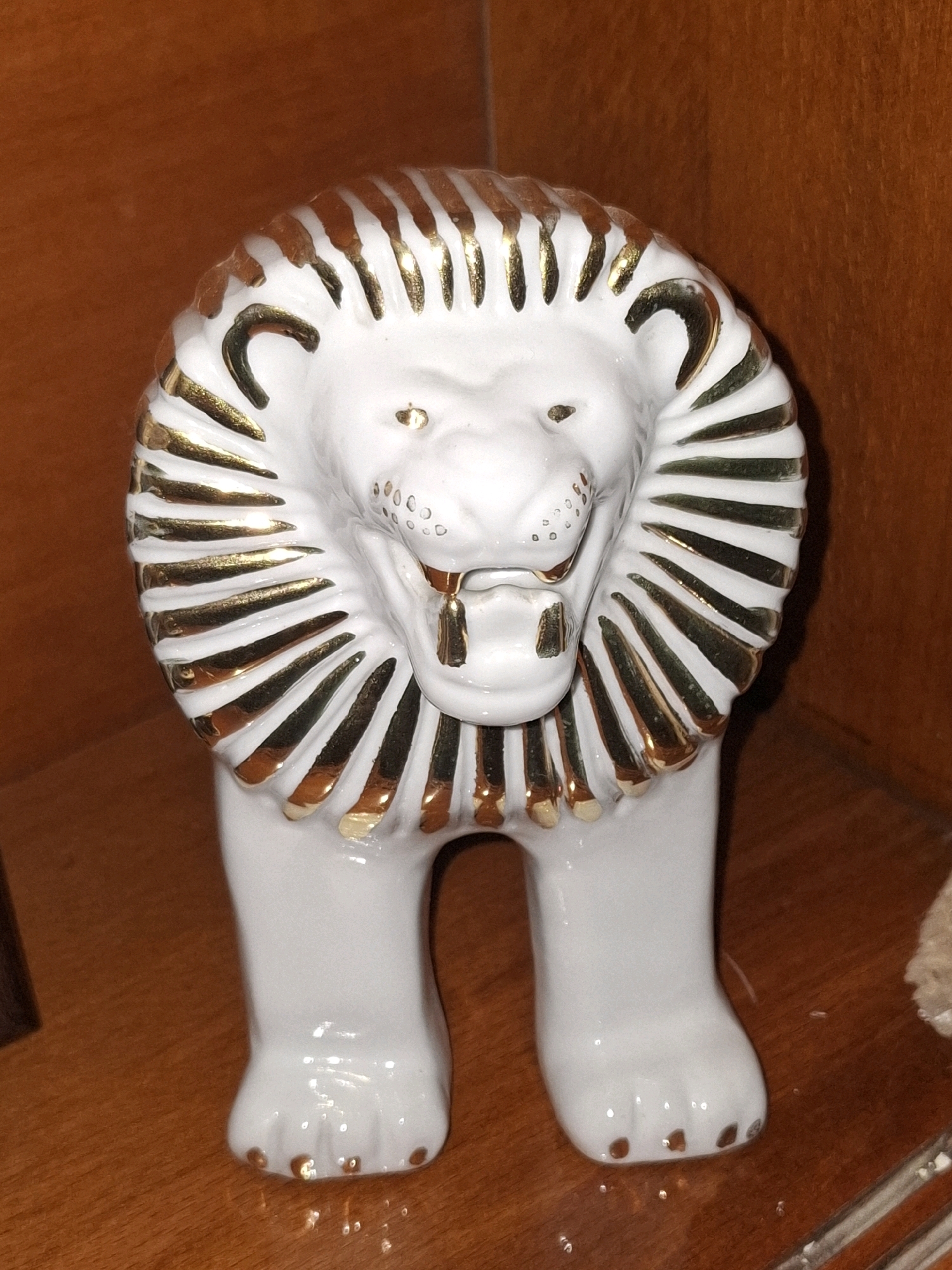 A white porcelain figurine of a lion is placed on a wooden surface. The lion has a mane with gold accents, painted eyes, and a slightly open mouth revealing gold accents on its teeth. The figurine stands on four legs, with gold accents on its paws. The background is a warm brown, likely a wooden shelf or wall. The lighting appears to be artificial and warm, casting soft shadows. The scene is static and indoors. The location in Almaty, Kazakhstan, is not discernible from the image itself.