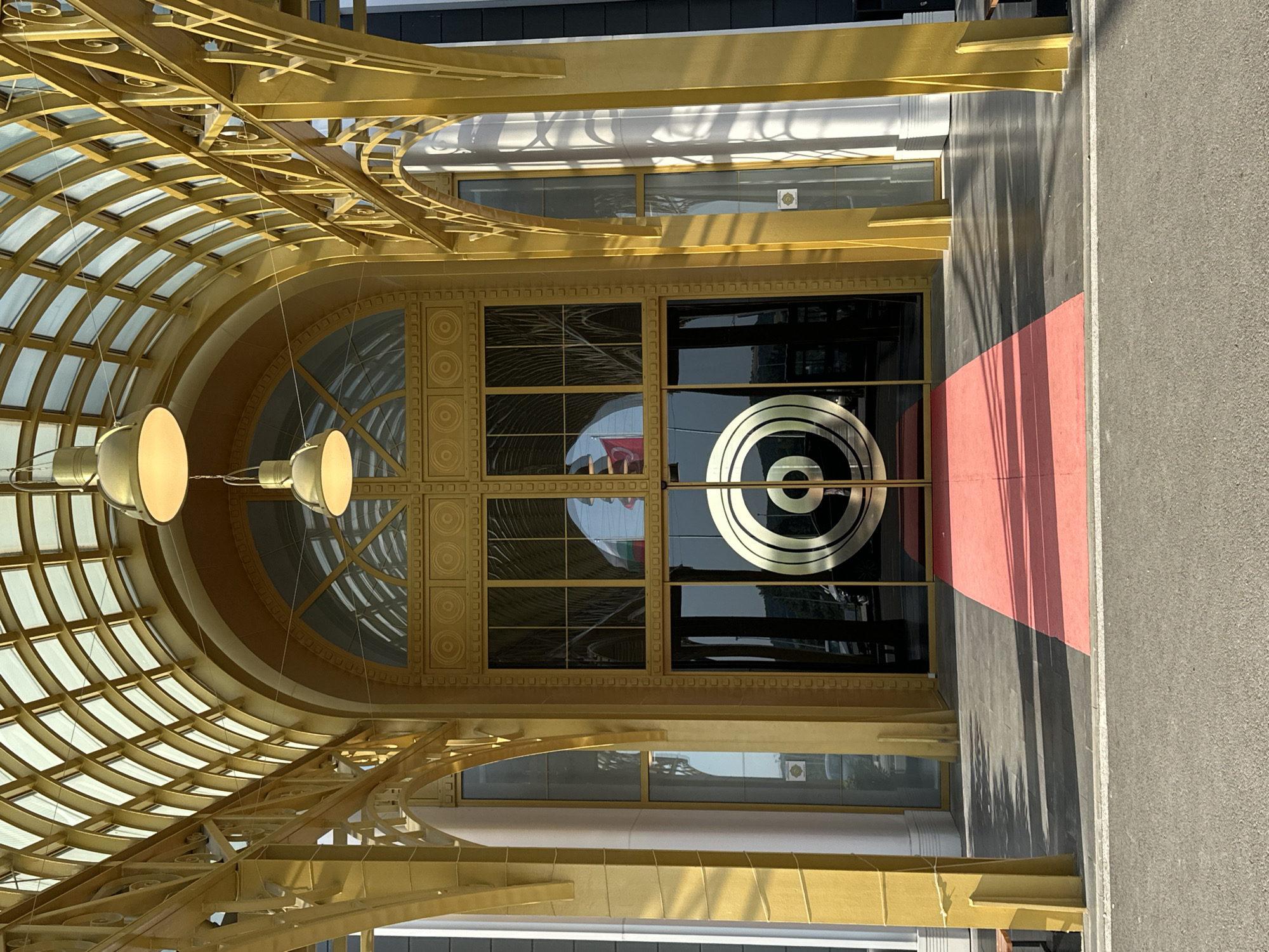 An ornate, golden architectural entrance stands prominently, featuring a grand arched canopy and a red carpet leading to the main doors. The structure, likely a commercial or public building in Kapitan Andreevo, Bulgaria, is illuminated by bright daylight, casting distinct shadows. The arched canopy overhead is composed of golden latticework and translucent panels, resembling a skylight. Two large, golden pendant lights hang from the canopy's ceiling. The facade is predominantly golden, with decorative elements, including circular motifs above the central doors and intricate scrollwork on the supporting arches and balustrades. The main entrance consists of double glass doors, dark and reflective, featuring a large, stylized golden concentric circle logo on the right door. Large arched windows above the doors and rectangular windows to the sides also have golden frames. Reflections in the glass show a landscape with hills and trees, vehicles, and a flag with horizontal red, white, and green stripes. A bright red carpet runner extends from the doors across a dark grey tiled pavement towards the foreground. A narrow asphalt road with a white line is visible at the very bottom of the image. No individuals are present in the immediate vicinity of the entrance.