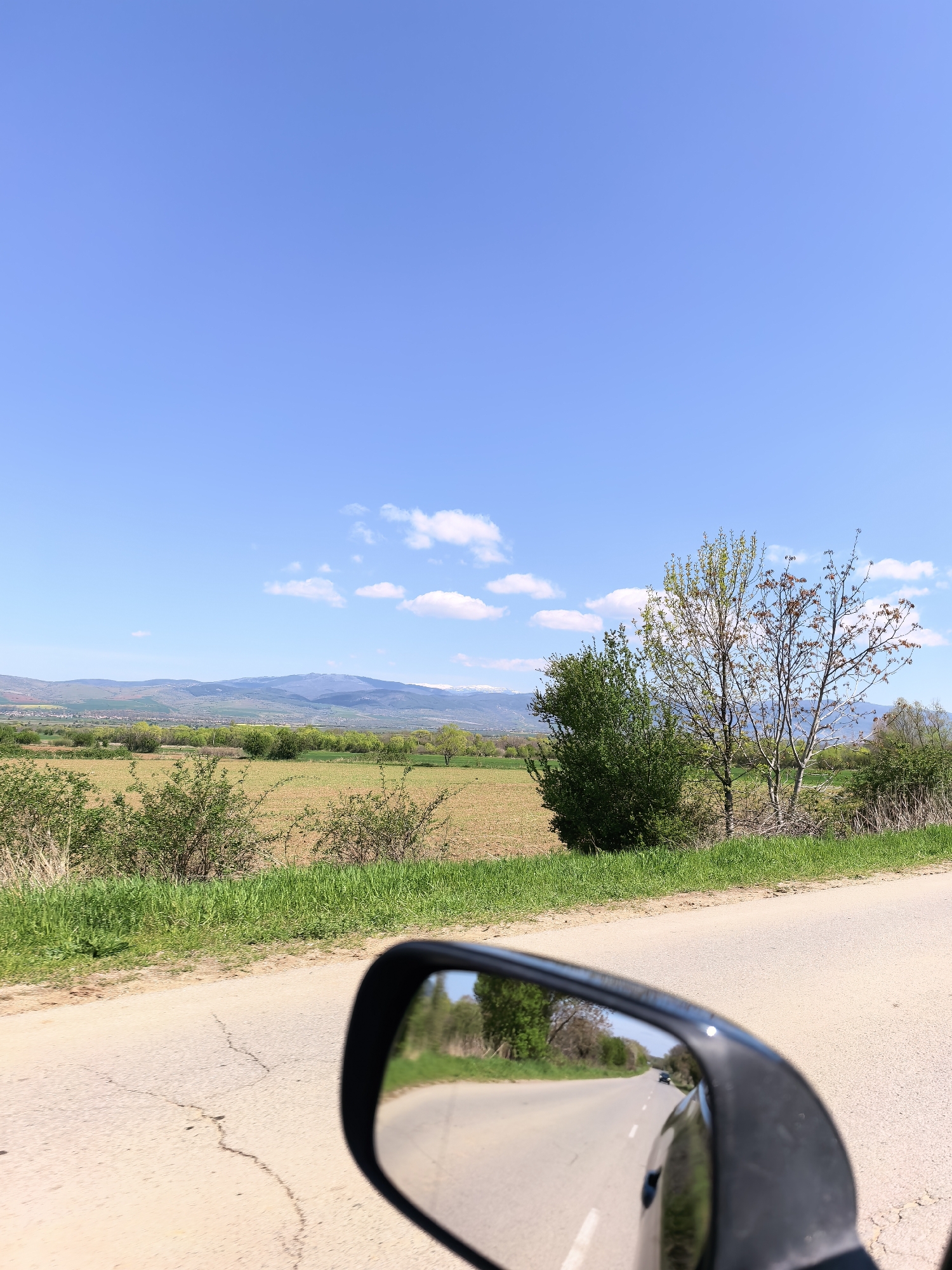 The image depicts a rural road scene under a clear blue sky with scattered clouds. In the foreground, a cracked asphalt road stretches into the distance. A car's side mirror reflects the receding road, lined with trees. The midground features a field with low-lying vegetation, bounded by taller trees and bushes. Distant mountains are visible on the horizon. The location is unknown.