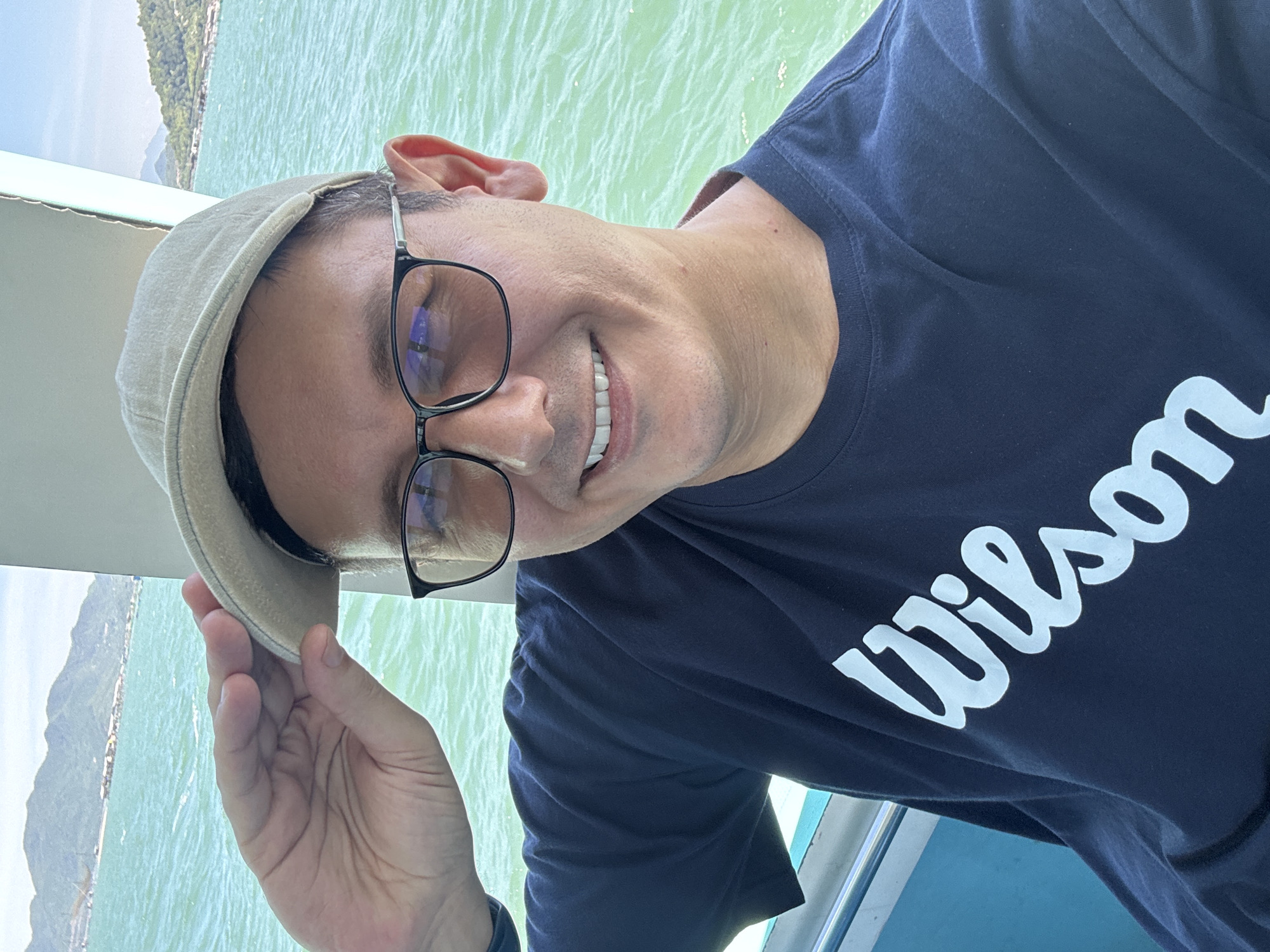 A man wearing a dark blue t-shirt with "Wilson" written in white, a beige baseball cap, and black-rimmed glasses is taking a selfie on a boat. He is holding the brim of his cap with his left hand. The man is smiling and looking at the camera. The background shows a body of turquoise water, a coastline with lush green hills, and some structures on the water. The scene appears to be in the vicinity of Bac Nha Trang Ward, Vietnam, given the tropical landscape and coastal setting.