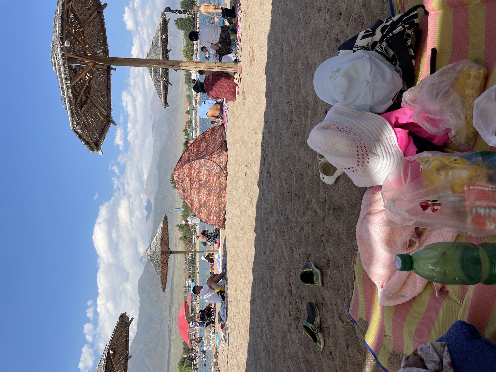 This image captures a scene on the sandy beach of Lake Issyk-Kul in Choktal, Kyrgyzstan. The foreground is dominated by a colorful striped mat laden with beach items, including a pink towel, a green plastic bottle, and various plastic bags filled with snacks. Two pairs of clogs are scattered on the sand nearby.
In the mid-ground, people are enjoying a sunny day. Several thatched umbrellas provide shade. A large, patterned tent is set up on the beach, and further back, people are swimming and relaxing in the shallow waters of the lake. The background features a clear blue sky with scattered clouds, and a range of mountainous terrain is visible in the distance, characteristic of the region. The overall atmosphere suggests a leisurely day at a popular beach resort.