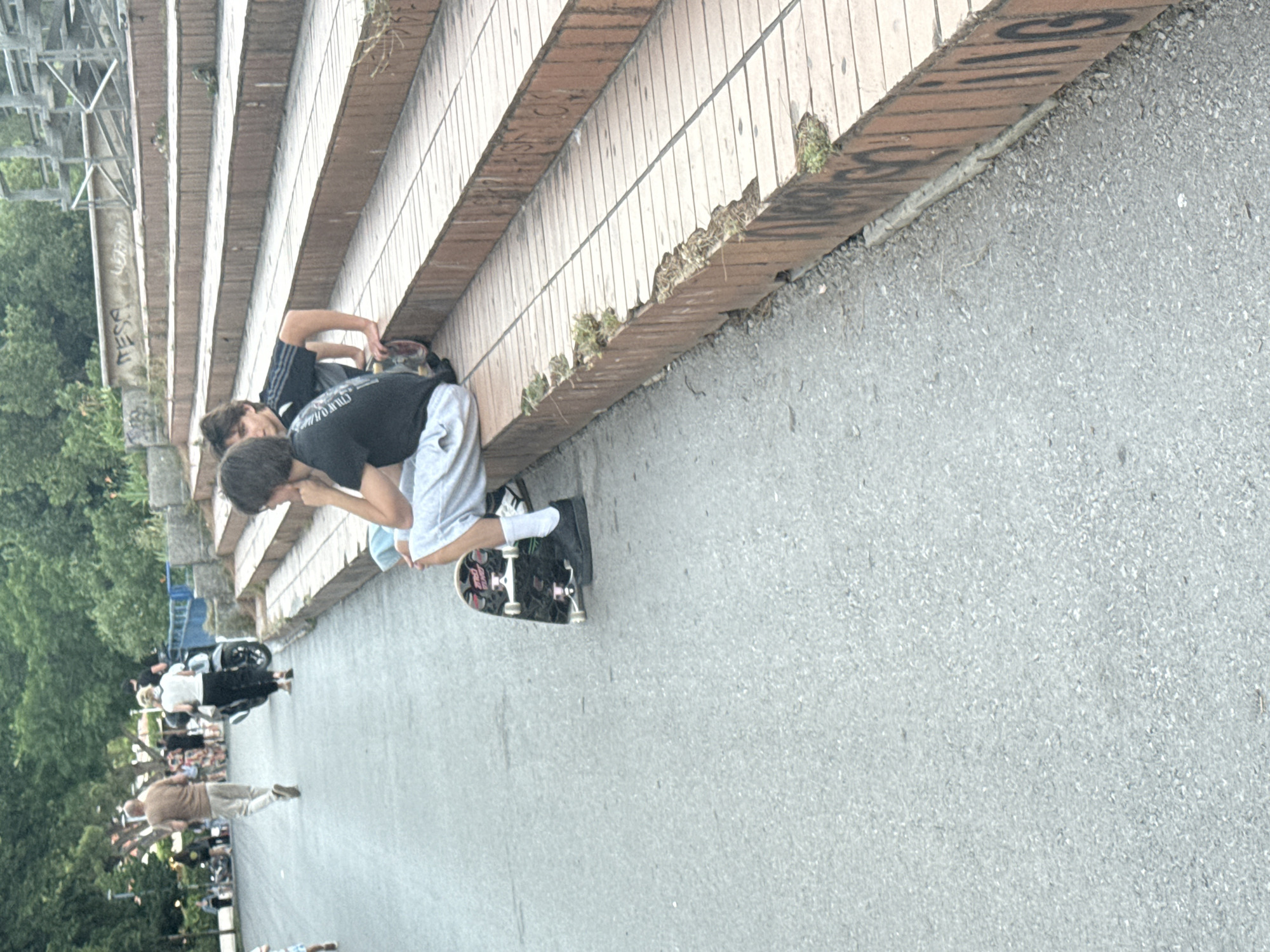 Two people are sitting on a skateboard at the base of a staircase in Budva, Montenegro. The staircase is made of brick and has graffiti on the side. Other people are walking in the background. The environment appears to be a public space with trees and a pedestrian walkway.