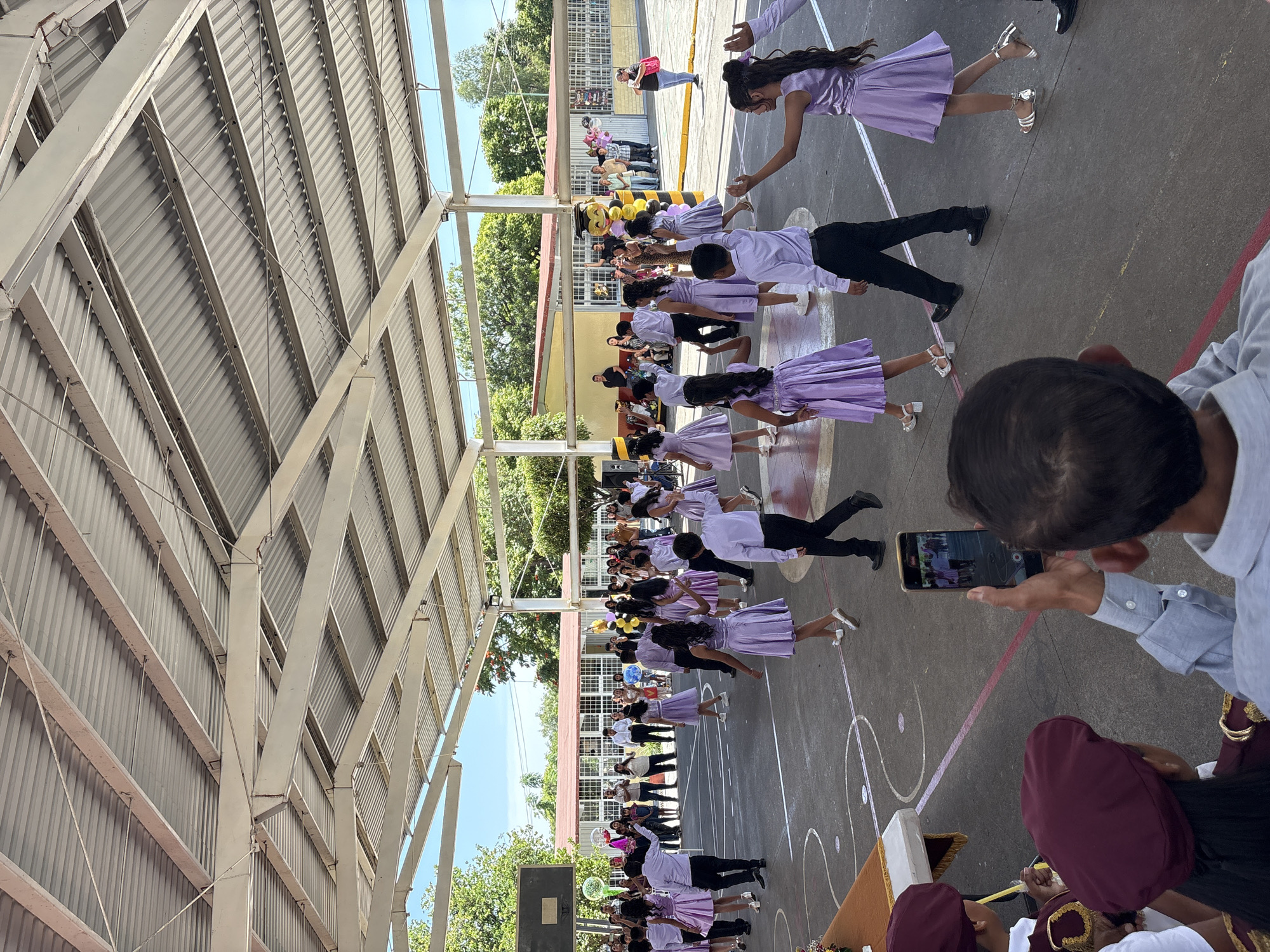 The image shows an outdoor event taking place under a covered pavilion with a corrugated metal roof supported by white metal beams. A basketball hoop is visible on the left. The ground is a dark asphalt court marked with white lines. Several children in matching purple dresses and white dress shirts and black pants are performing a dance. Some adults are watching the performance from the sidelines, some of whom are taking pictures with their phones. A large mascot, resembling a bee with a graduation cap, is standing near the center. Balloons and streamers add to the festive atmosphere. The background reveals a building with red-tiled roofs and trees. This scene is taking place at an unspecified location in Tejalpa, Mexico.