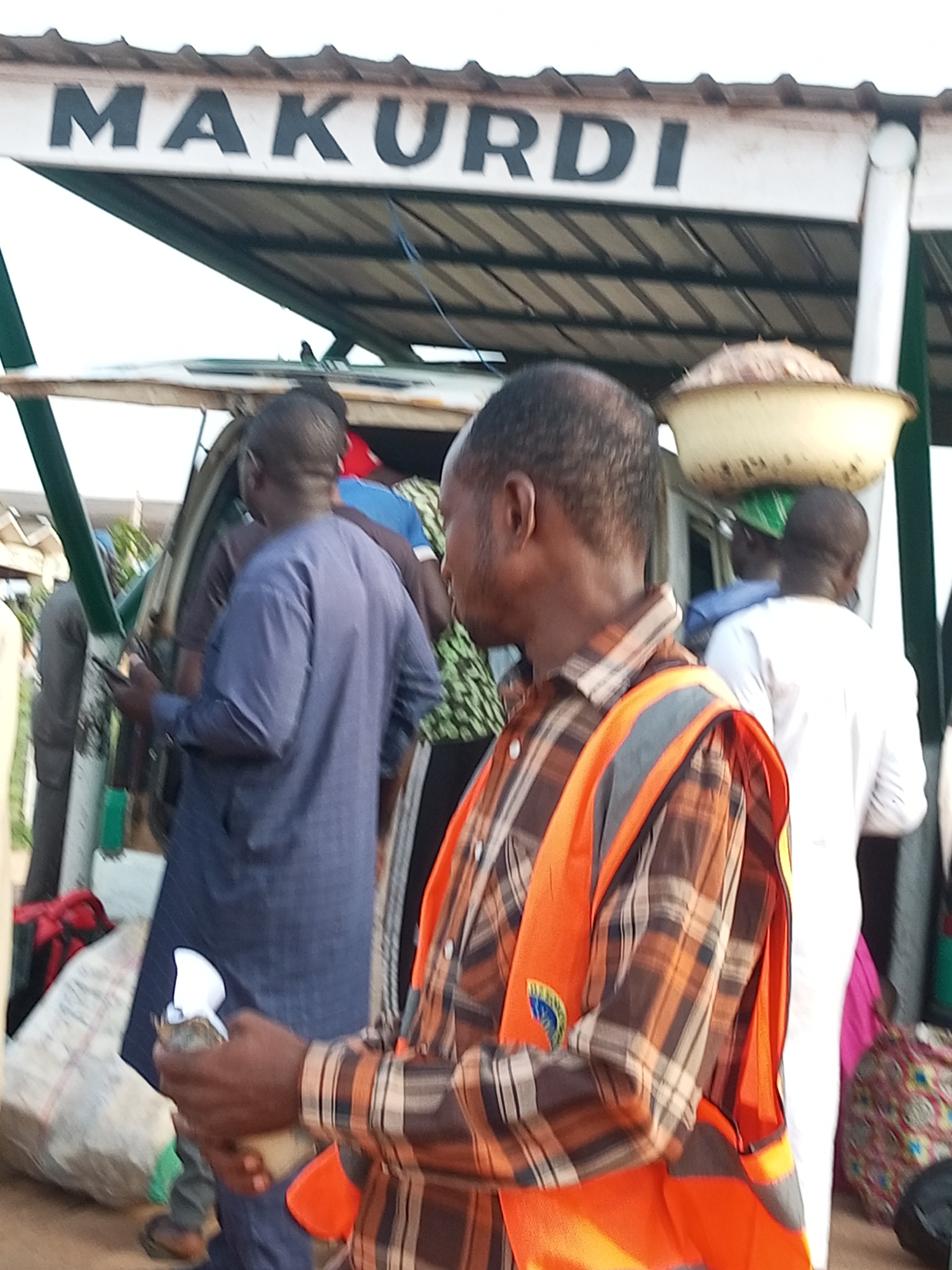 A man wearing a plaid shirt and an orange safety vest is holding a white object and what appears to be money. He is standing near a vehicle. In the background, a sign reads "MAKURDI". The scene appears to be outdoors, possibly at a transportation hub or market. The general setting suggests an urban environment within Nigeria.