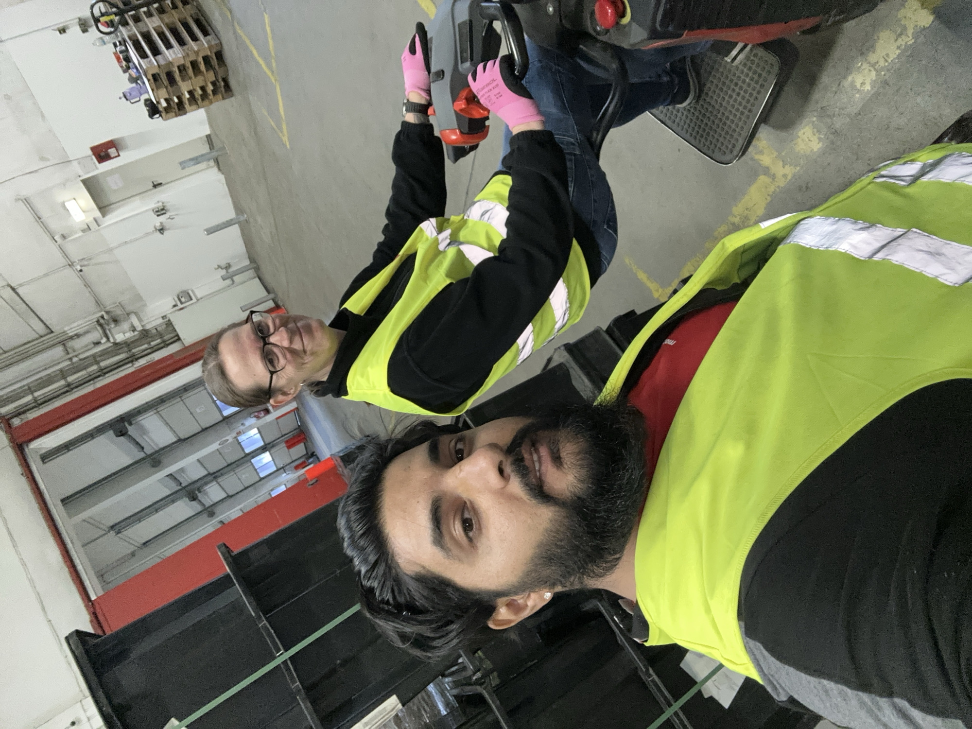 Two individuals are present in what appears to be a warehouse or distribution center. The person in the foreground is a man with dark hair and a beard, wearing a bright yellow safety vest over a red shirt. The person behind and to the right is a woman with glasses, also wearing a yellow safety vest over a black shirt. She is operating a powered pallet jack, as indicated by her hand position on the controls and her feet on the operator platform. The environment is characterized by industrial shelving units on the left, a concrete floor with yellow markings, and large open doorways in the background. A stack of wooden pallets is visible in the middle ground. The lighting suggests an indoor industrial setting. Based on the context provided, this scene is located in Schweitenkirchen, Germany.