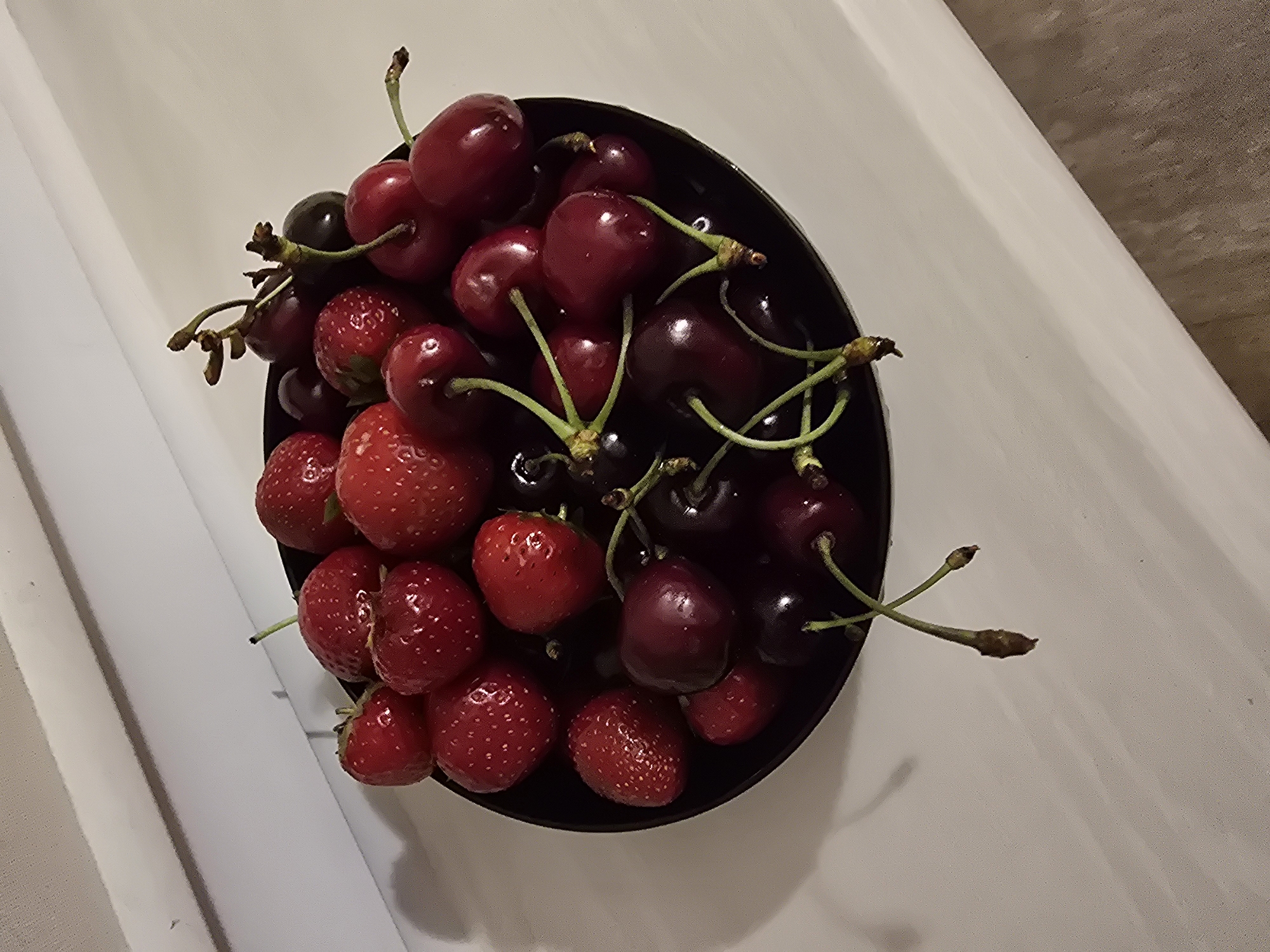 A black bowl of strawberries and cherries is placed on a white surface, with a white wall in the background. The strawberries are red with visible seeds, and the cherries are a deep red color. Some of the cherries have stems attached. The lighting suggests an indoor setting. The image is taken from a slightly elevated angle. There is no indication of Sofiivska Borshchahivka, Ukraine.