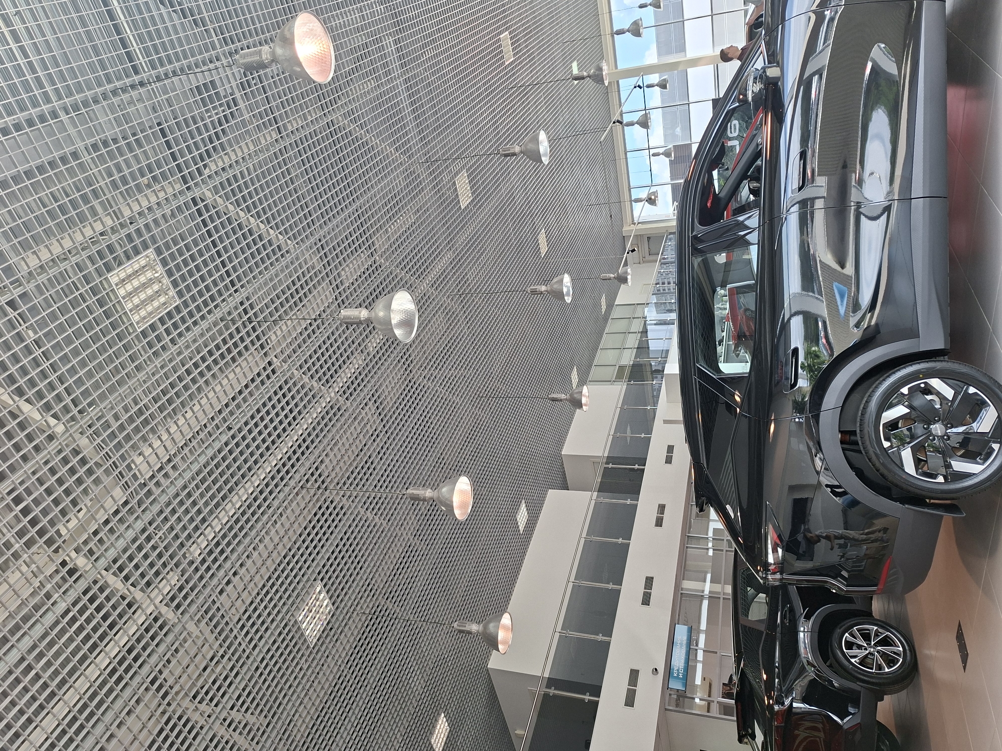 The image depicts the interior of a modern car dealership in Izhevsk, Russia. The scene is dominated by a black SUV parked prominently in the foreground, with another black SUV partially visible to its left. The dealership features a high, grid-like metal ceiling with numerous pendant lights. Large windows line the back wall, offering a view of the sky and distant buildings. A few people are present, including one man standing near the right side of the frame, seemingly observing the cars. A sign with Russian text, likely indicating services like "CREDIT AND INSURANCE" (КРЕДИТОВАНИЕ И СТРАХОВАНИЕ), is visible on a glass partition. The overall environment suggests a clean, well-lit, and contemporary commercial space.