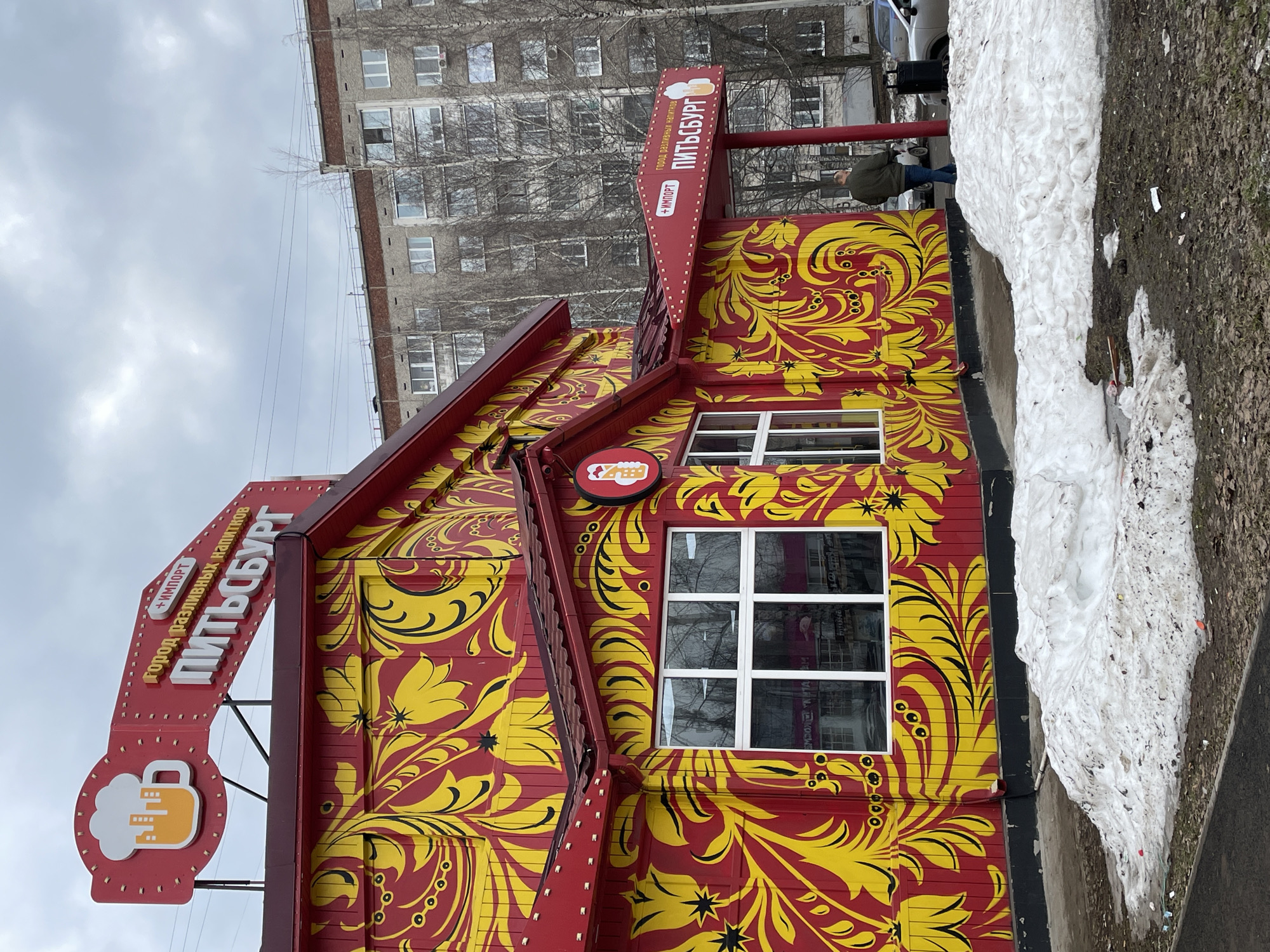 The image shows a colorful building in Izhevsk, Russia, decorated in a traditional Khokhloma style with red and gold floral patterns. It's identified as "ПИТЬСБУРГ" (Pit'sburg), a beer establishment, as indicated by the signs and the beer mug symbol. A snowdrift is present on the right side of the image, indicating winter or early spring. A person is partially visible near the building, and an apartment building is visible in the background under a cloudy sky.