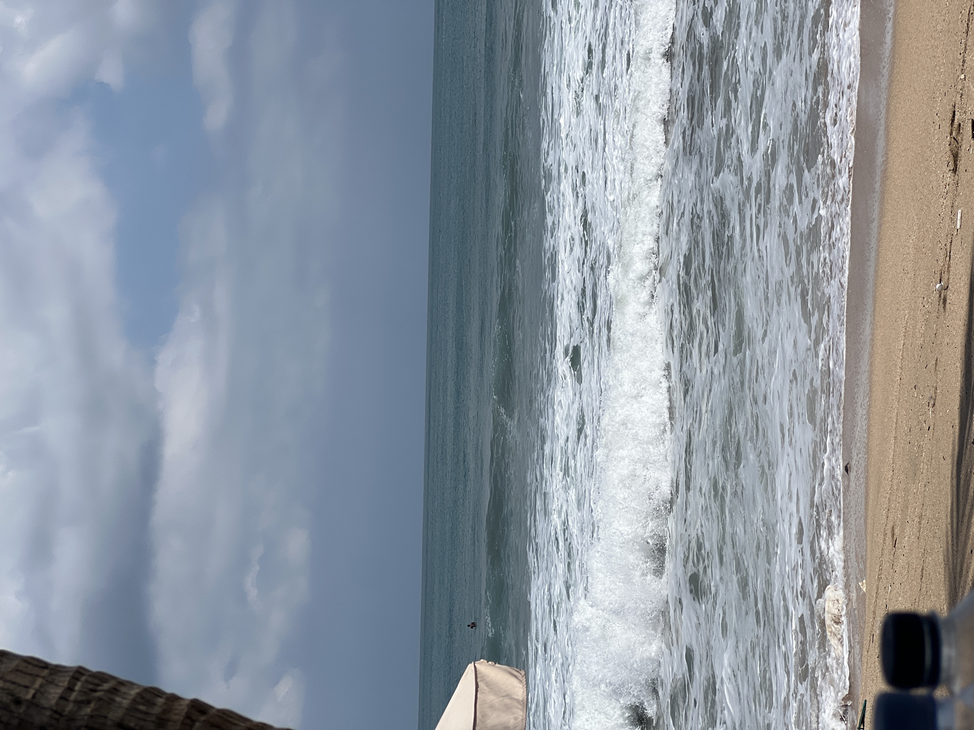 A medium-shot, eye-level, vertical view depicts a stormy, overcast sky above the turquoise waters of Kuta Beach, Indonesia. The ocean is active, with white-capped waves breaking near the sandy shore, where debris is scattered. In the middle distance, a solitary surfer rides a wave, while to the left, a portion of a beige beach umbrella is visible, partially obscuring the view. The foreground is dominated by the sand and the tops of two water bottles.