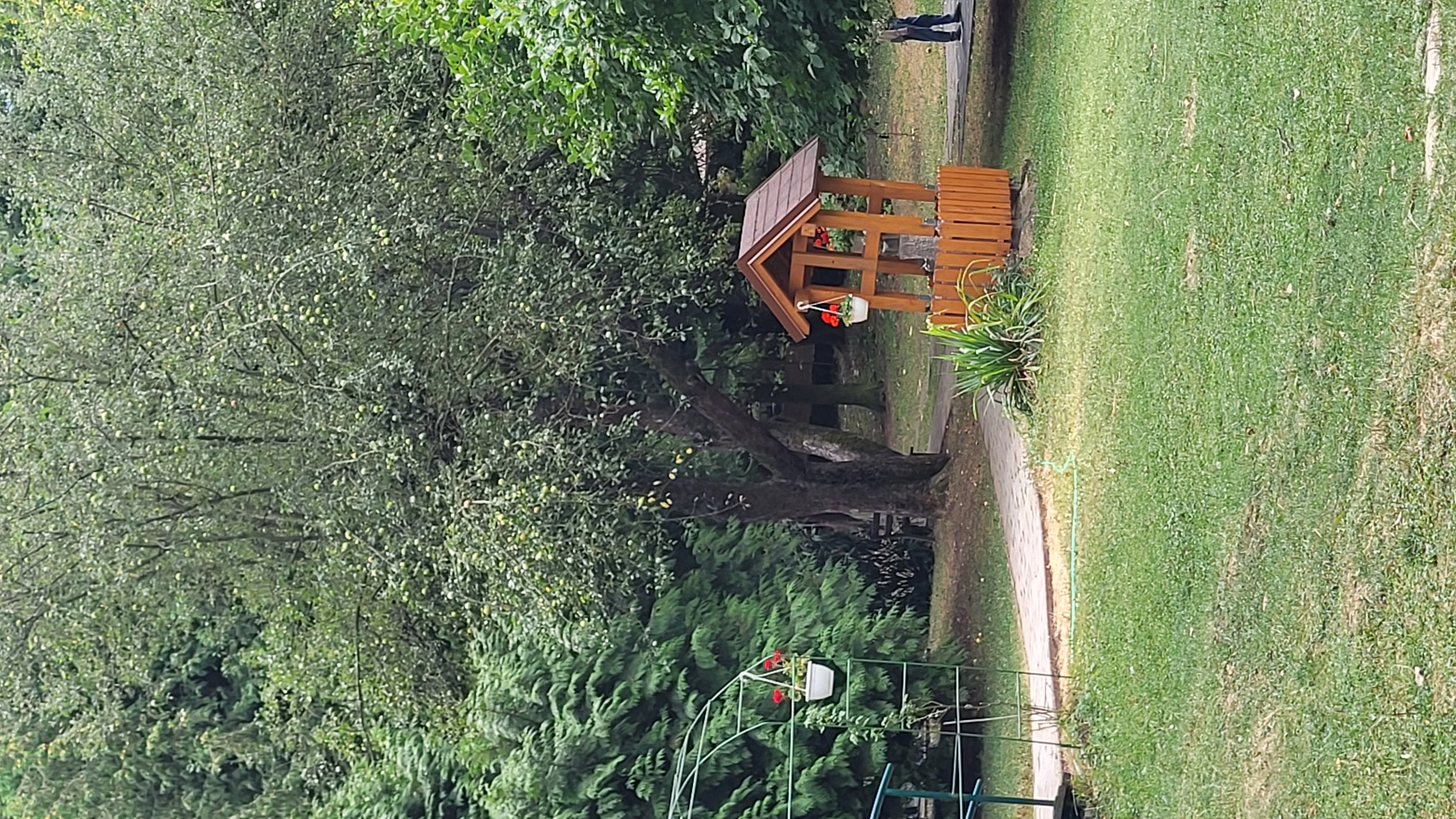 The image depicts a well with a wooden roof and base, decorated with a hanging flower basket. A paved pathway winds through a grassy area, bordered by trees and foliage. To the left, a green metal archway with a hanging flower basket is visible. In the background, a person walks along the pathway. The scene suggests a park or garden setting. This image was taken in the area of the Slavic Library in Prešov, Slovakia.