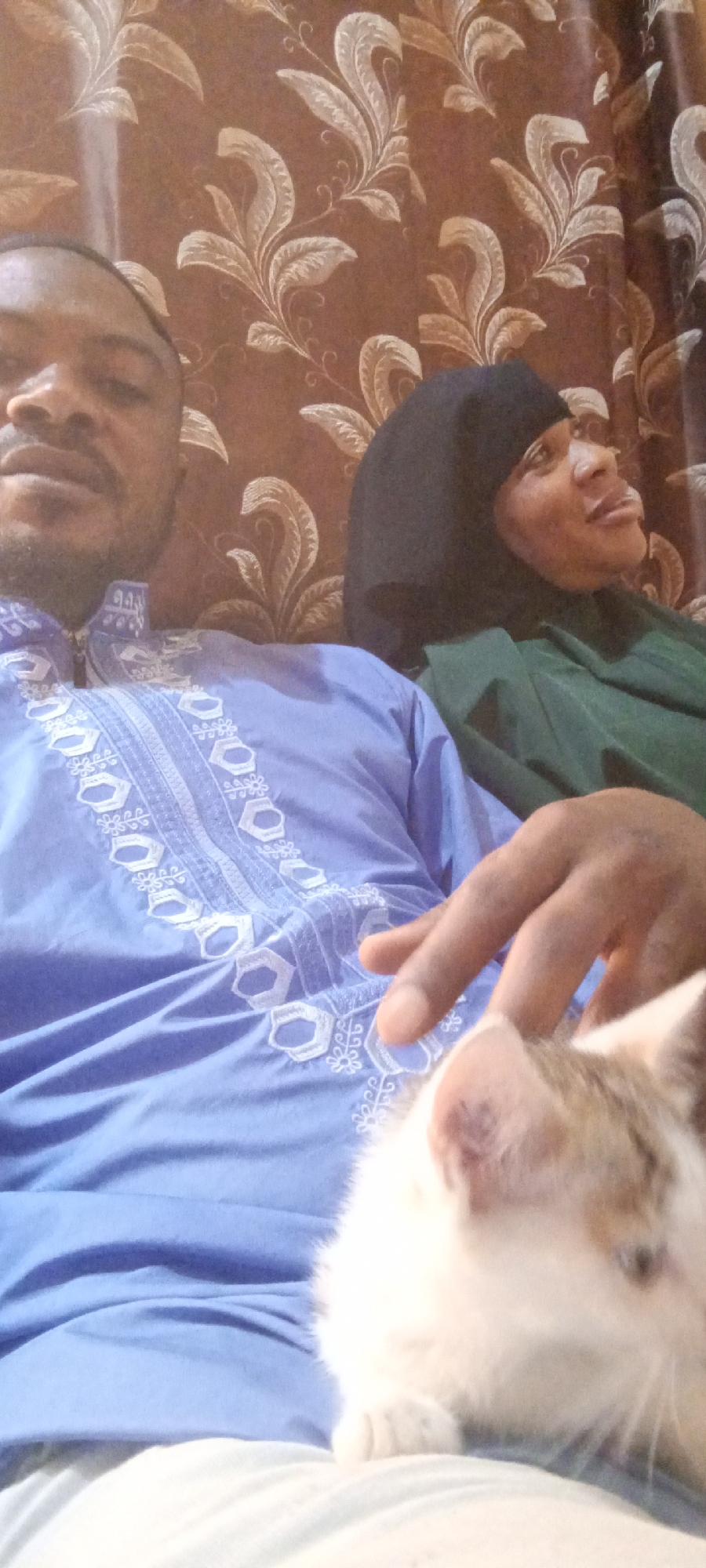 The image depicts two individuals and a cat indoors. A dark-skinned man is seated wearing a blue traditional embroidered shirt. Next to him is a woman wearing a black hijab and a green garment. A white and light brown cat is resting in front of the man. The backdrop consists of brown patterned wallpaper. The location is Gama, Nigeria, though the city name is not visually discernible in the photograph. The specific actions and interactions between the people and the cat are not clear.