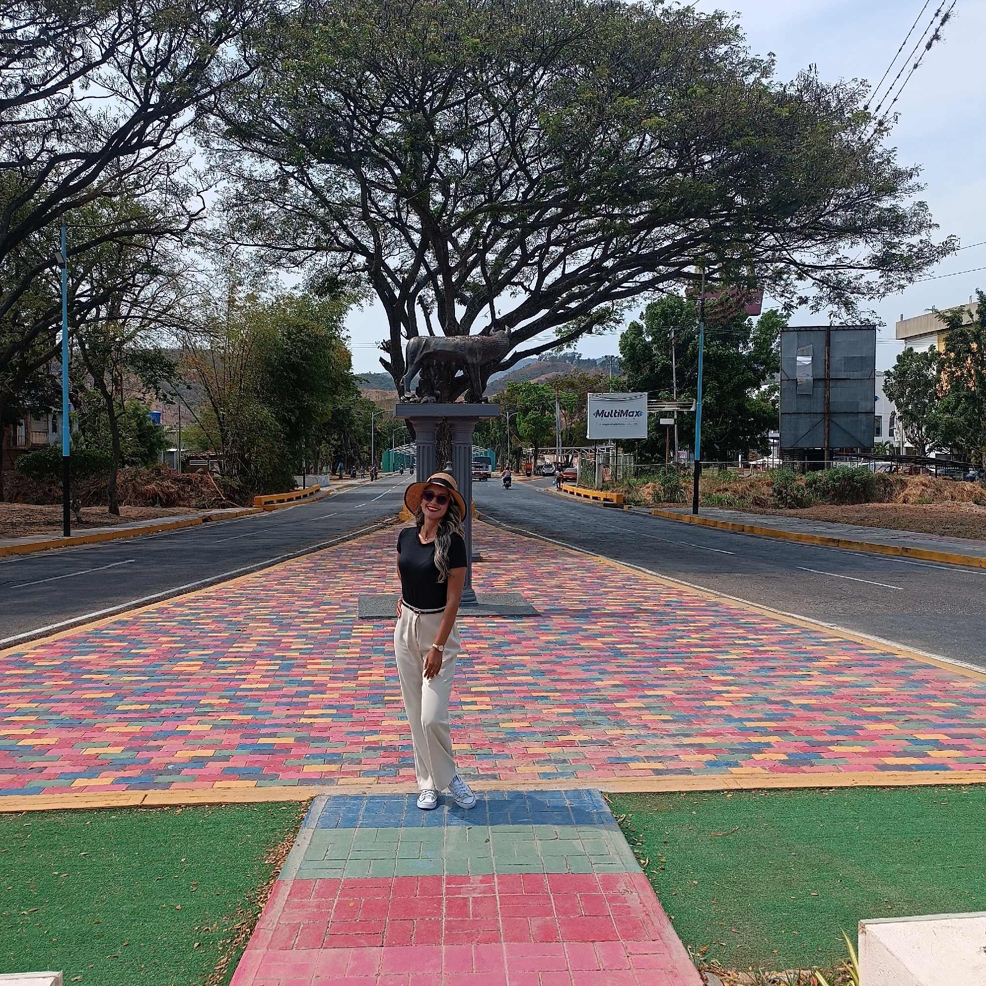 A daytime photograph depicts an urban scene on a divided avenue in La Victoria, Venezuela, featuring a central median.
In the foreground, a light-skinned adult female stands centrally on a colorful paved surface. She wears a black short-sleeved top, light-colored high-waisted trousers, white sneakers, a straw hat, and sunglasses. Her left arm is relaxed, and her right hand rests on her hip. The paved surface beneath her consists of rectangular blocks painted in red, blue, yellow, and green hues, arranged in a brick pattern. To her immediate sides, artificial green turf is visible.
Behind the woman, the colorful paving extends to a two-lane asphalt road on each side, divided by a wider median. The roads have white lane markings. Further back on the central median, a bronze-colored statue of the Capitoline Wolf (Lupa Capitolina), depicting a wolf suckling two infants, is mounted on a dark pedestal composed of two columns. Immediately behind the statue, a very large, mature tree with a wide, dark trunk and extensive leafy canopy dominates the midground.
To the right of the median, a white "MultiMax" billboard is visible, along with another large, empty billboard structure. Along the roadsides, various trees and green foliage are present. Piles of what appear to be organic debris or brush are visible on the unpaved verges. A blue street light pole stands on the left side of the median.
In the background, sparse vehicle and pedestrian traffic can be observed on the roads. Distant hills or mountains covered in vegetation frame the horizon under a light blue sky with scattered clouds.