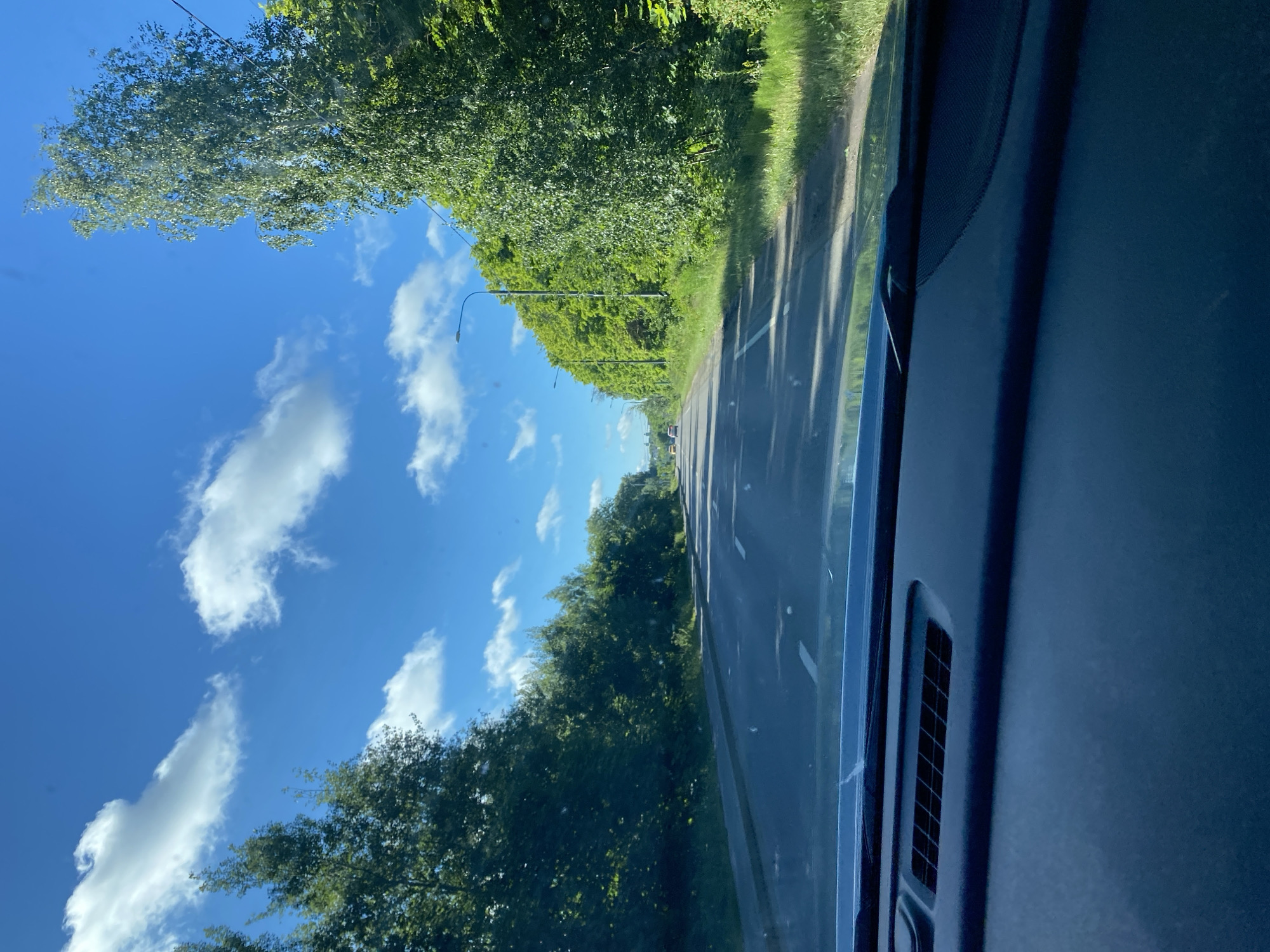 The image depicts a view from inside a vehicle looking forward along a paved road lined with lush green trees and vegetation on both sides. The sky is a bright blue with scattered white clouds. There are a few cars visible in the distance on the road. The foreground shows the dashboard of the vehicle. The scene appears to be a suburban or rural road on a sunny day.