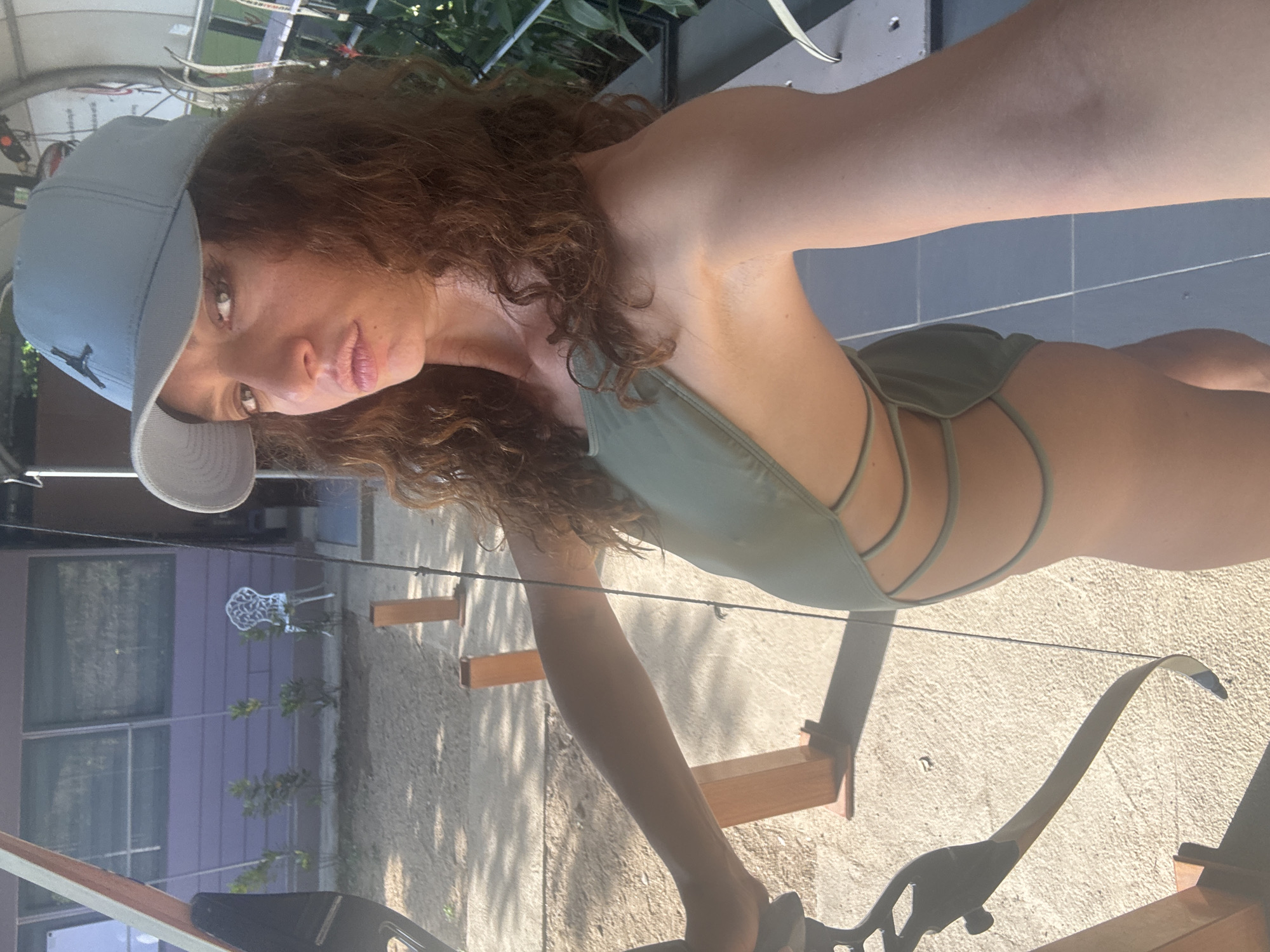 A woman wearing a light-colored baseball cap and a green swimsuit is holding a bow and arrow. She is outdoors on a concrete surface. Behind her is a building with purple siding and visible windows. A white chair is in the background, partially obscured by vegetation. The location is not discernible as being in Đà Nẵng, Vietnam.