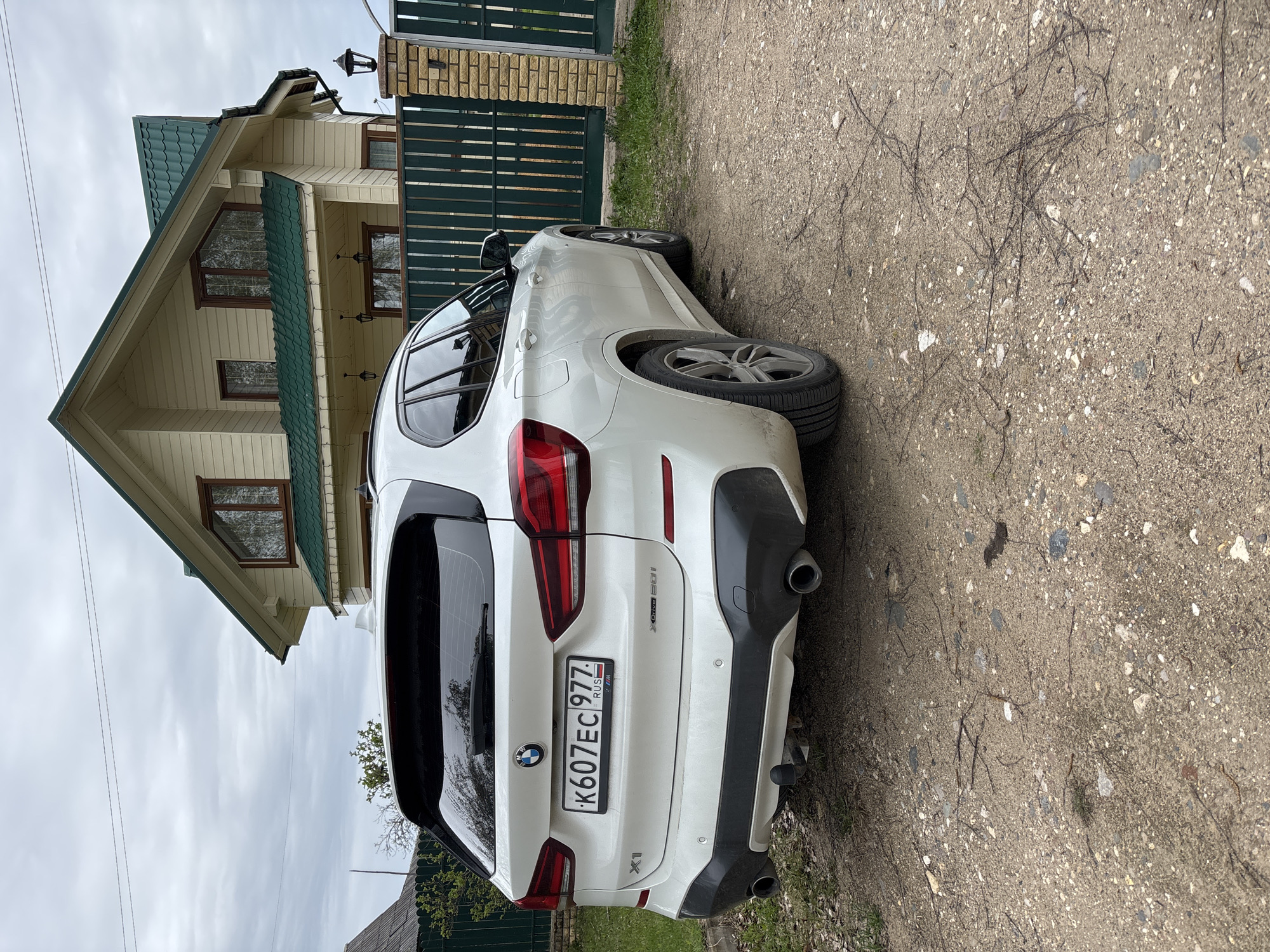 A white BMW X1 with license plate K607EC777RUS is parked on a dirt road next to a residential building. The building has a light yellow exterior with green roofing and dark-framed windows. A green metal fence runs along the property line. The location is Zaprudnya, Russia, as indicated by the vehicle registration plate.