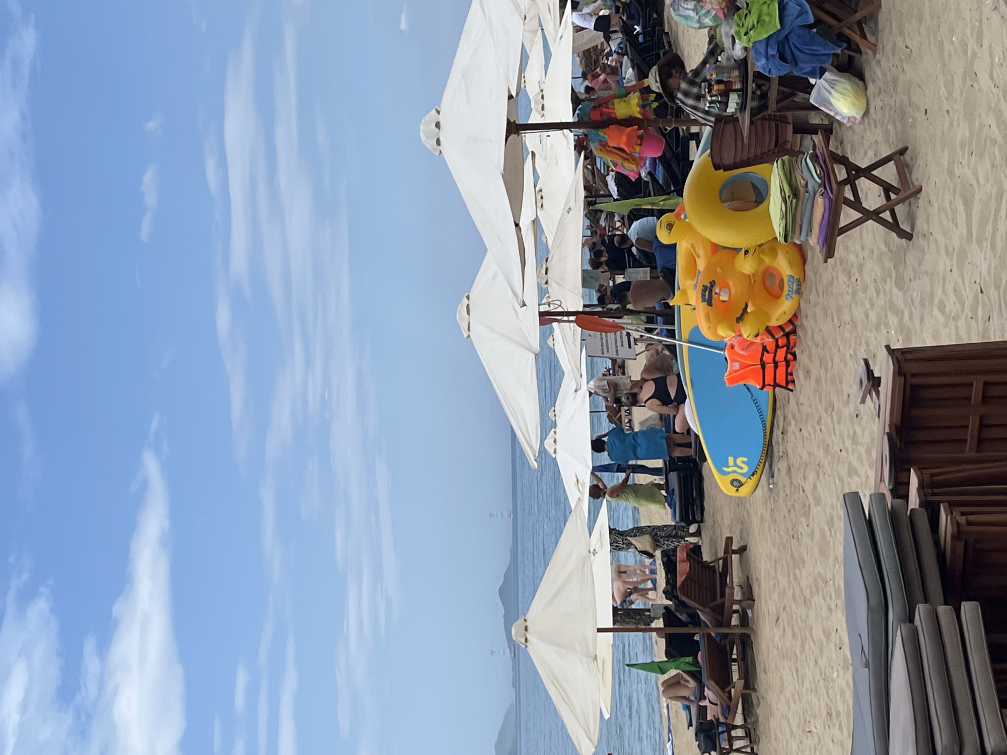 A sunny day unfolds on a sandy beach in Nha Trang, Vietnam, with the turquoise ocean meeting a bright blue sky dotted with white clouds. White umbrellas stand tall, providing shade to beachgoers lounging on chairs and towels. In the foreground, colorful inflatable toys, including a duck and a large yellow ring, lie on the sand, alongside a blue surfboard and orange life vests. People are scattered across the beach, some in groups, others individually, enjoying the seaside. Some are standing, interacting with each other, while others are seated or lying down. The water is calm, with distant islands visible on the horizon.