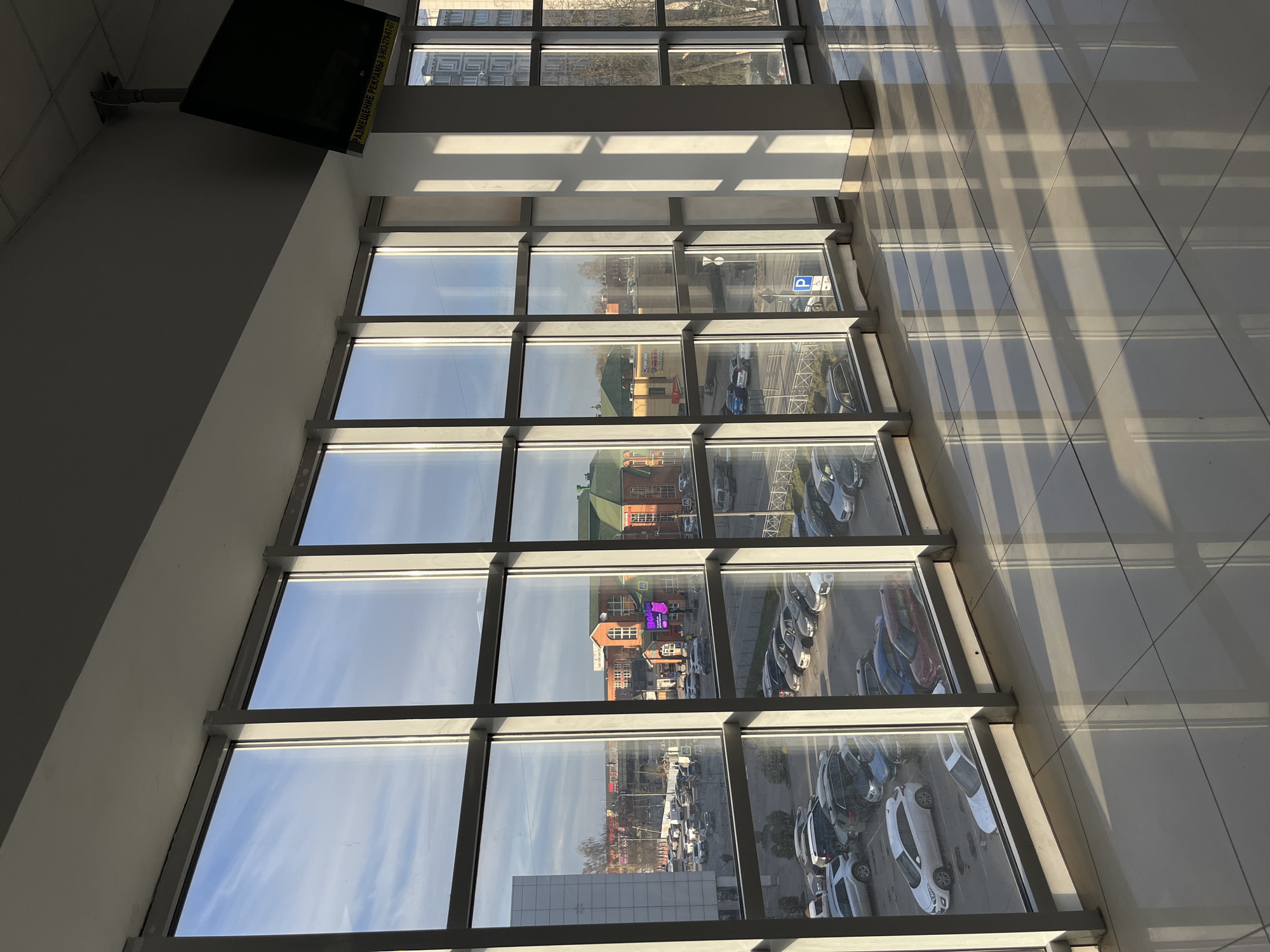 The photograph depicts an interior view through a multi-paned window overlooking a street and parking area in Klin, Russia. Visible outside the window are buildings, cars parked in a lot, street signs including a parking symbol, and general urban scenery under a partly cloudy sky. Inside, the window's frame casts shadows on the tiled floor. A screen, possibly a television, is mounted on the upper left wall. No people or actions are visible in the image.