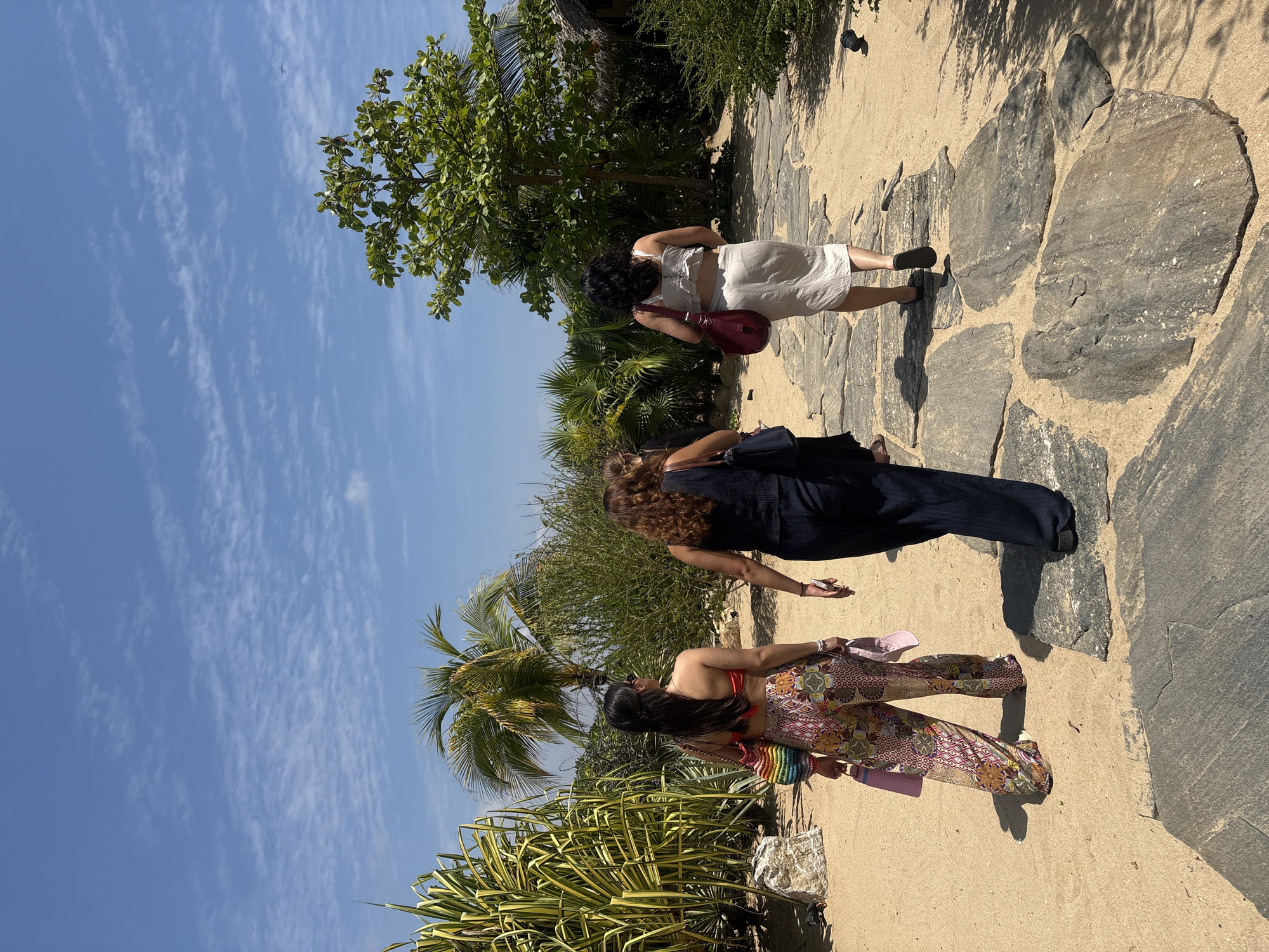 The image captures three women walking along a stone pathway in an outdoor setting. The pathway is made of large, irregularly shaped stones embedded in sand. A blue sky with wispy clouds is visible above. On the left side of the pathway, there is tropical vegetation, including palm trees and shrubs. The woman furthest from the camera wears a white dress and carries a red bag. The woman in the middle wears a dark jumpsuit. The closest woman wears floral pants and a red top. Ministerios de Amor Eterno Asociación Cultural, Mexico cannot be determined by the image alone.