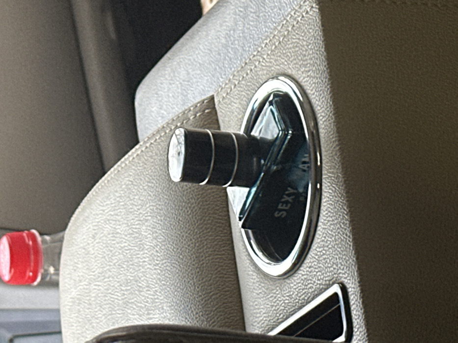 The scene depicts a detailed view inside a vehicle, located in Huangdao District, China. A rectangular glass perfume bottle with a dark, cylindrical cap rests within a chrome-rimmed cup holder. The bottle, which appears to contain a light blue liquid, prominently displays the word "SEXY" and partial text "AM" on its side. The cap is dark, possibly black, and features three distinct horizontal silver bands. The cup holder is integrated into a padded center console or armrest, upholstered in a light beige or gray material with a finely textured surface. In the blurred background, a transparent plastic bottle with a red screw-top cap is partially visible, positioned behind the console. No individuals are present in the image, and no active interactions are occurring.