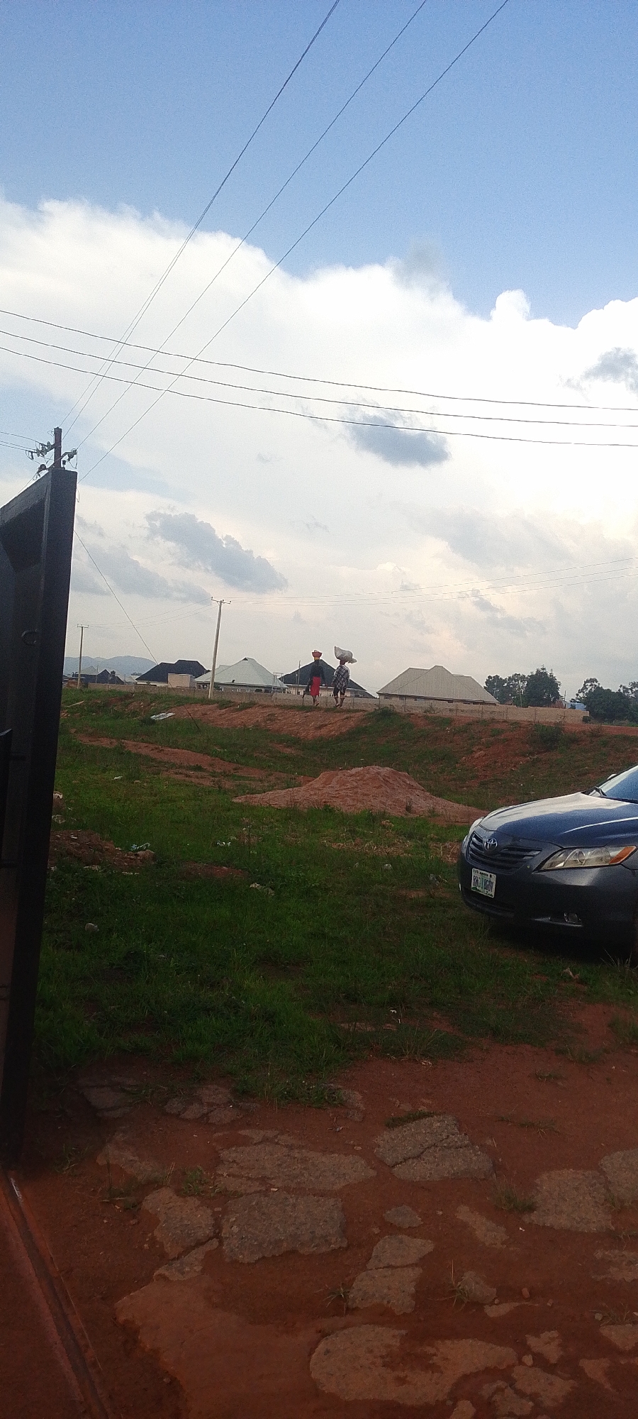 The image captures an outdoor scene in Jos, Nigeria, under a cloudy sky. In the foreground, a dark metal gate is visible on the left, and a portion of a dark sedan is parked on the right. The ground is a mix of reddish-brown earth and patches of cracked asphalt in the immediate foreground, transitioning to uneven grassy terrain further back.
In the mid-ground, two individuals are walking away from the camera. The person on the left wears a red hat and a dark garment, while the person on the right carries a white bag on their head and wears a checkered garment. They appear to be walking along a raised dirt path. Behind them, several unfinished or newly constructed houses with light-colored roofs are spread across the landscape. Utility poles and overhead power lines crisscross the sky, indicating the presence of electrical infrastructure.
The scene suggests a developing residential area. The sky is filled with scattered clouds, hinting at a potentially overcast or partially sunny day. The overall impression is one of everyday life and construction in a rural or semi-urban setting. No visible text is present in the image.