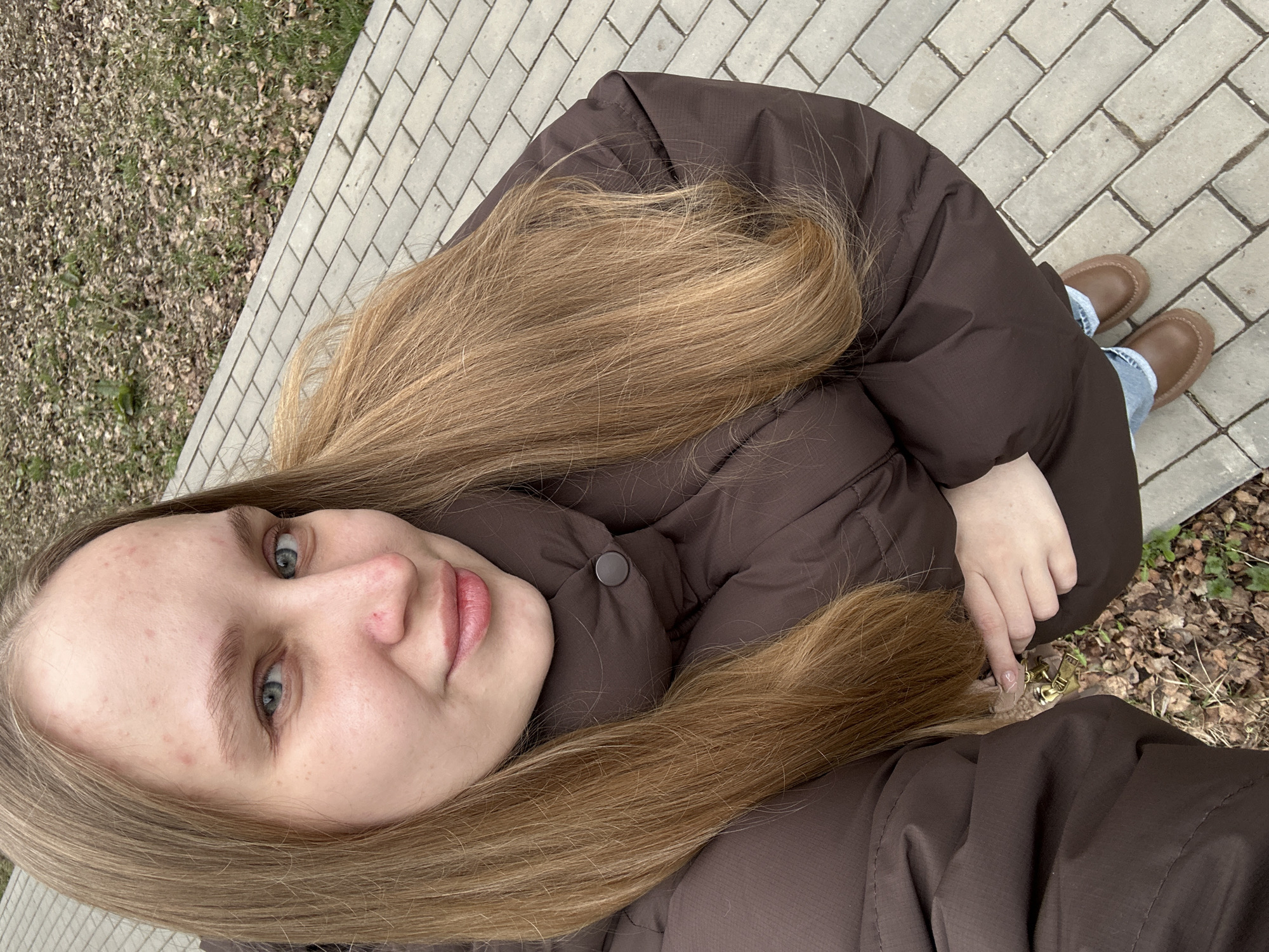 A selfie-style photograph shows a young woman with long, light-brown hair, wearing a dark-brown puffer jacket, tan-colored shoes, and light-blue jeans. Her face is visible, with light skin and blue eyes. The background consists of a paved sidewalk with a brick pattern, and to the left, an area of soil, leaves, and grass. The location could be a park or a pathway alongside a street, although the specific area of Chaikovsky, Russia, is difficult to ascertain from this single image.