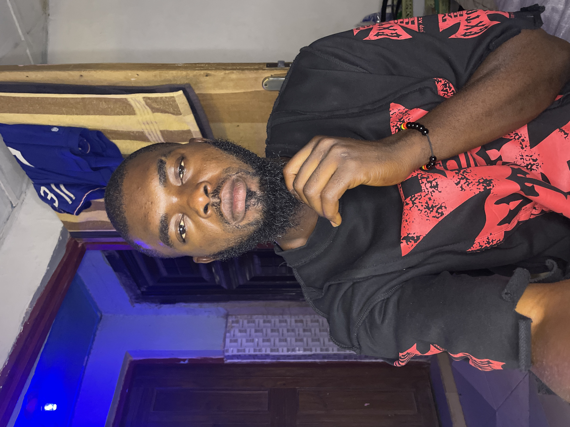 An eye-level indoor portrait captures an adult male, likely in his 20s or 30s, with dark skin, a full black beard, mustache, and short dark hair, situated in a domestic environment within the city of Jos, Nigeria. He wears a black long-sleeved crew-neck shirt featuring distressed collar and cuff edges, adorned with irregular red graphic designs across the fabric. A black beaded bracelet with intermittent red, yellow, and green beads is visible on his left wrist. His right hand is raised, with fingers curled, resting against his chin as he looks directly into the camera with a neutral, steady gaze.
The background is dimly lit with mixed lighting. A bright blue LED bulb, visible in the upper left corner, casts a strong blue hue over a light-colored wall and the top of a dark reddish-brown wooden door frame. A dark wooden panel door is partially visible on the far left. Adjacent to this, a section of light grey tiles with a geometric pattern is present. In the upper right background, a blue garment (possibly a shirt with white numbering like "31" or "JE") hangs from above, next to a vertically draped, striped towel in shades of tan, brown, and black. A light-colored wall continues to the right, behind which a silver-colored door handle mechanism is visible on a doorway or partition. The lower right foreground shows indistinct items resting on a surface.