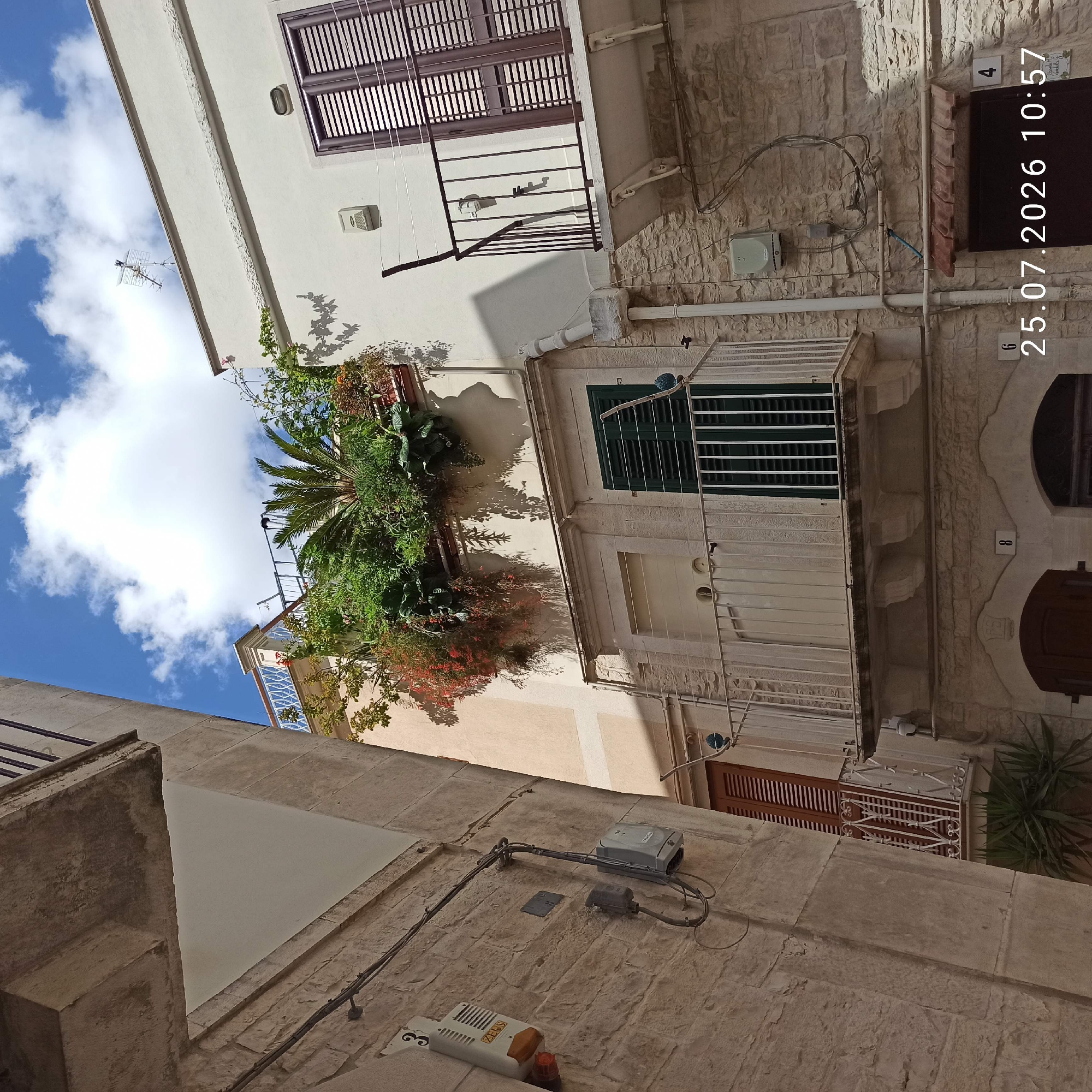 The image depicts a narrow street in Ruvo di Puglia, Italy, characterized by a building with stone walls and white plaster. The architecture features multiple balconies, some adorned with lush greenery and flowering plants, creating a vertical garden effect. One balcony in the foreground has a white metal railing, while another above it features a dark green shuttered window. A brown shuttered window is also visible on the left side of the frame. Electrical conduits, boxes, and an antenna are mounted on the building's exterior. The scene is bathed in sunlight, casting shadows from the plants and architectural elements onto the walls. The sky is blue with scattered white clouds. A timestamp overlay in the bottom right corner reads "25.07.2026 10:57". There are no people visible in the image.