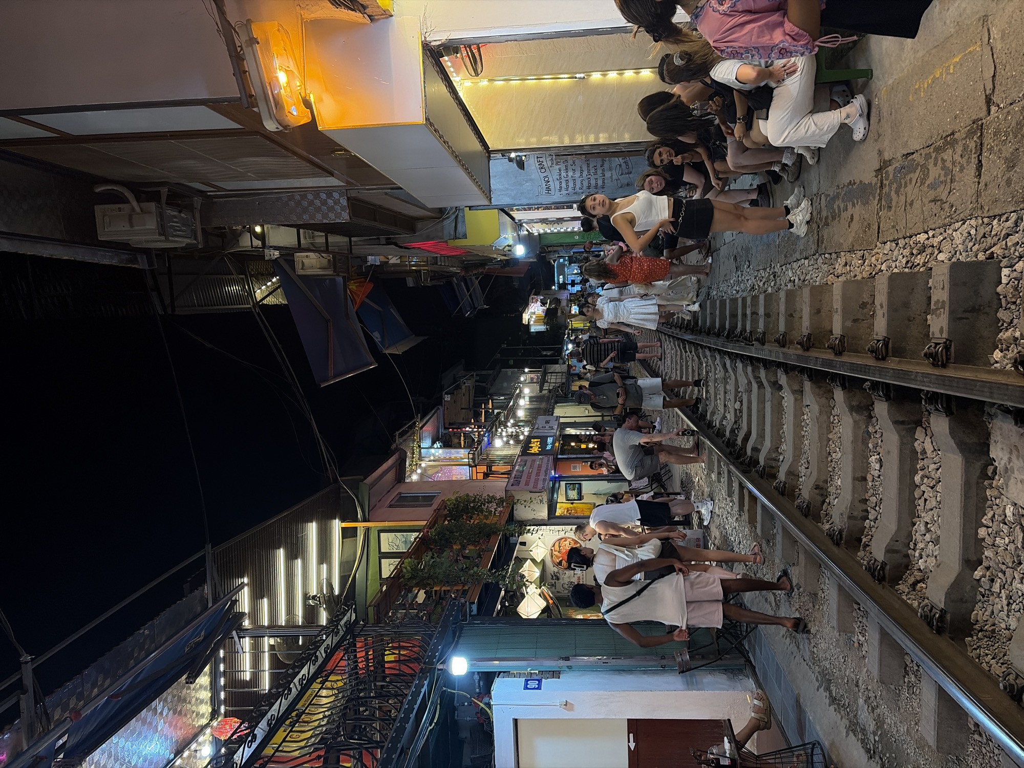 A night scene on Hanoi's Train Street, a narrow urban alleyway with active railway tracks serving as a thoroughfare and seating area for outdoor cafes. People, dressed casually in t-shirts, shorts, and dresses, are seated on benches and chairs on either side of the tracks, illuminated by warm, ambient lighting from the surrounding buildings and overhead string lights. Some individuals are standing, observing the surroundings or interacting with each other. The railway tracks, made of concrete sleepers and gravel, are a prominent feature in the foreground, disappearing into the distance. The buildings flanking the street appear to be a mix of residential and commercial establishments, with signs and storefronts visible. The overall atmosphere is one of relaxed evening social activity in an unusual urban setting.